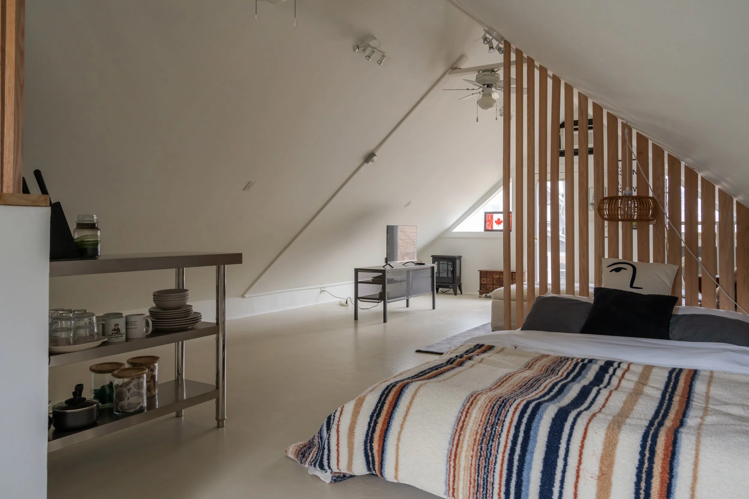 A minimalist attic bedroom with a sloped ceiling, a bed covered with a striped blanket and pillows, a wooden partition, a small TV on a stand, and a window with a Canadian flag sticker. The room features modern lighting fixtures and a neutral color p