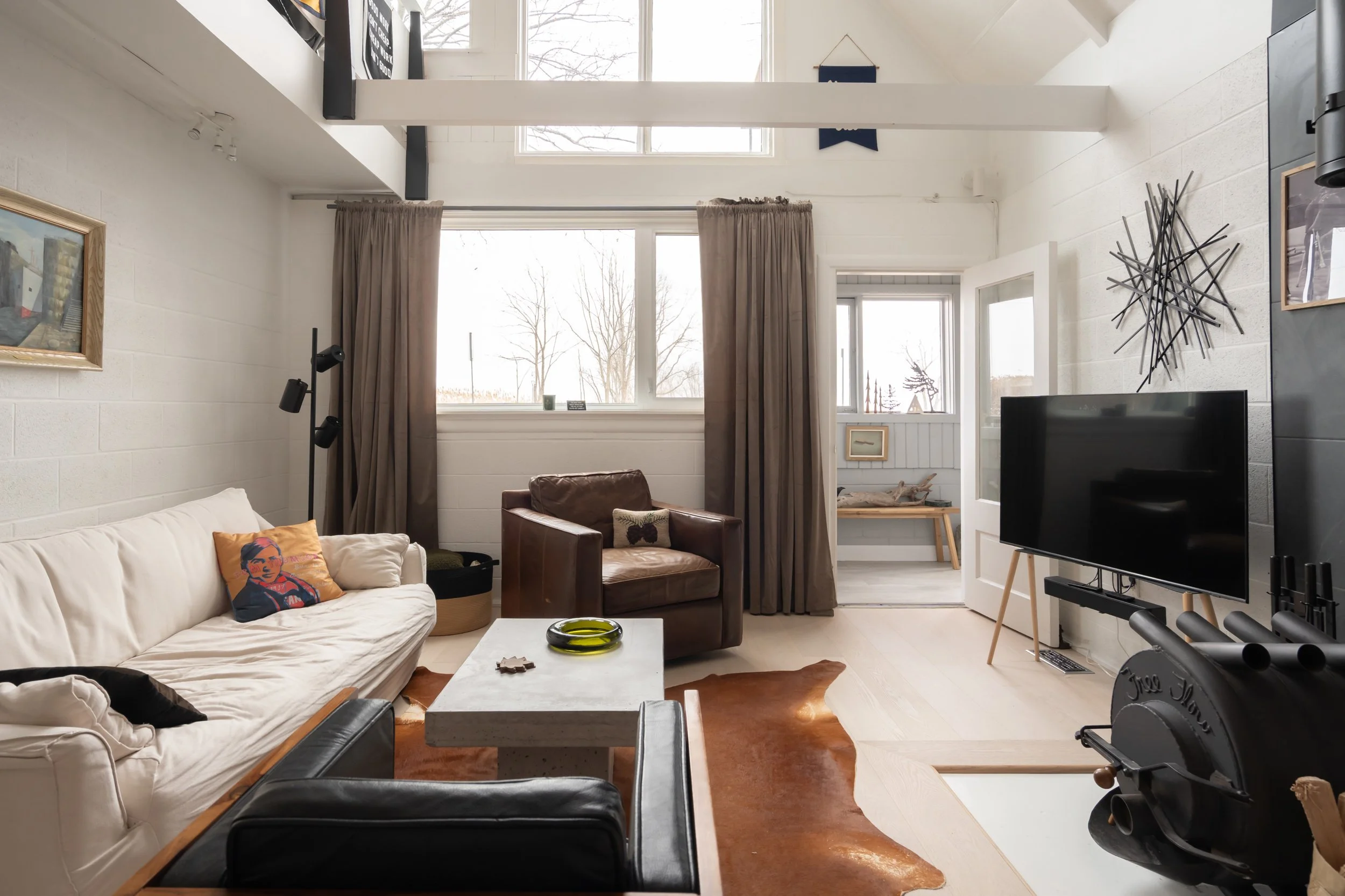 Living room with beige sofa, brown leather armchair, black chair, and a white coffee table on a brown cowhide rug. Large window with beige curtains, a flat-screen TV, and modern wall art. Light-colored walls and flooring, with a door leading to a bri
