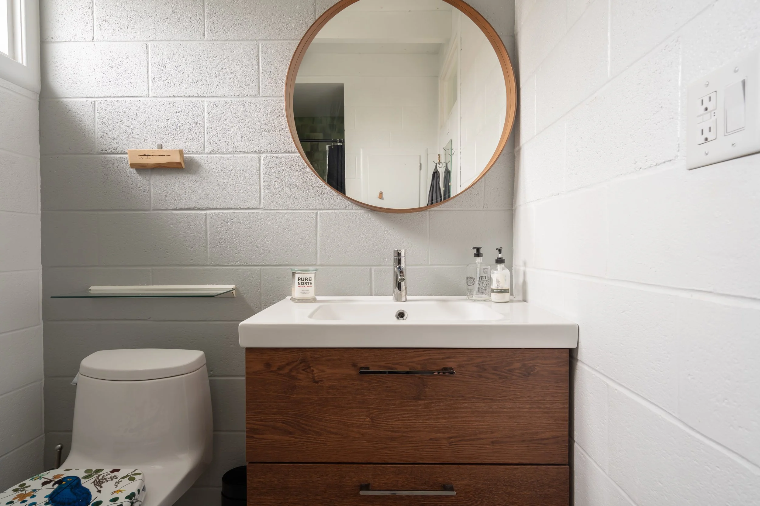 A modern bathroom with white painted brick walls, a wooden framed round mirror above a white countertop sink with a wooden cabinet below. To the left, a white toilet with a floral seat cover, a small wall shelf, and a glass on the sink. There are bot