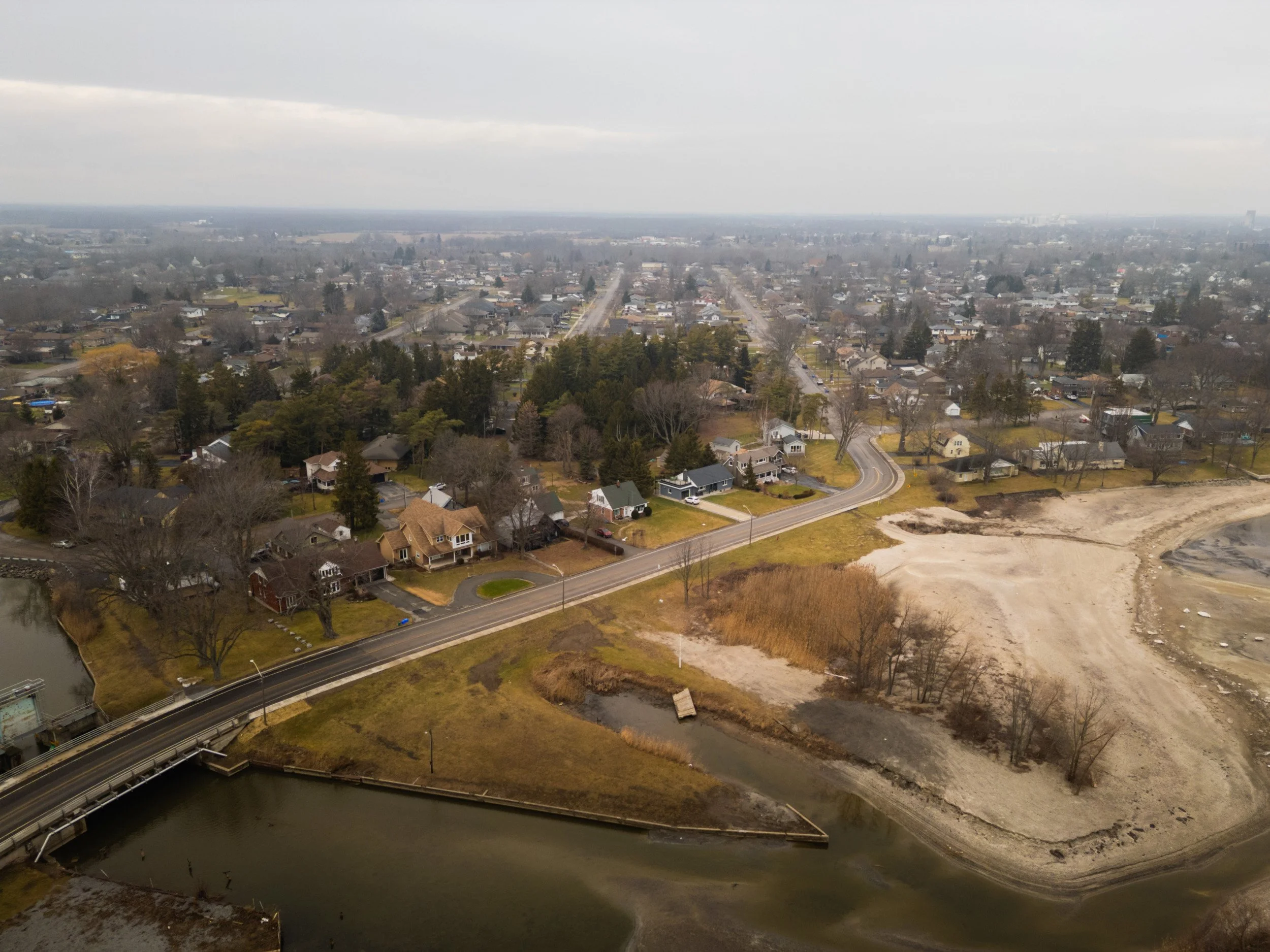 Aerial view of a suburban neighborhood with houses, a bridge over a body of water, and a large empty construction site on the right, under a cloudy sky.