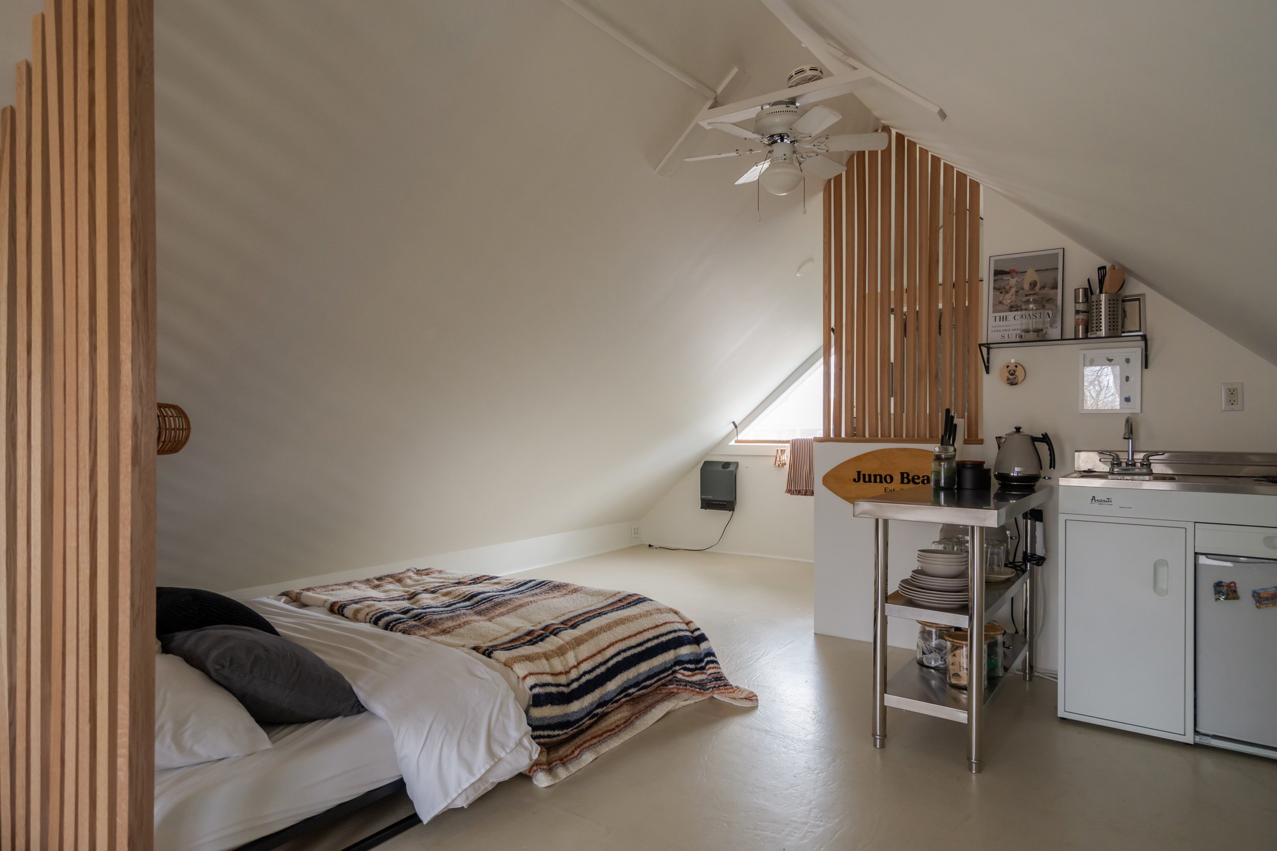 A small attic room with a bed on the left, a window with blinds, and a kitchenette with a sink, a microwave, a coffee maker, and shelves on the right.