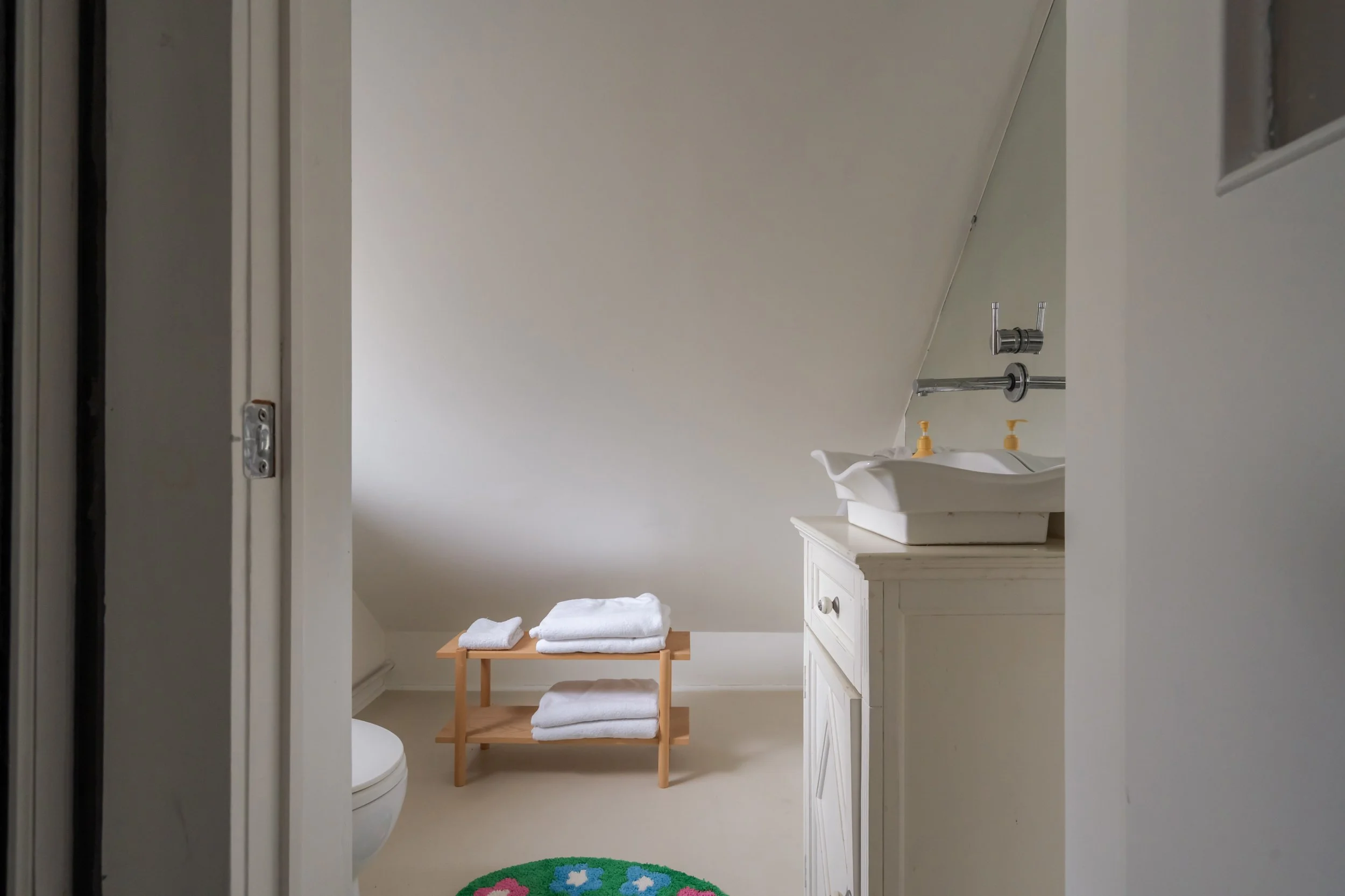 A small bathroom with a white vanity, a sink with yellow soap dispensers, a mirror above, a white toilet, a wooden towel rack with folded white towels, a green circular rug with colorful flower patterns, and an angled ceiling.