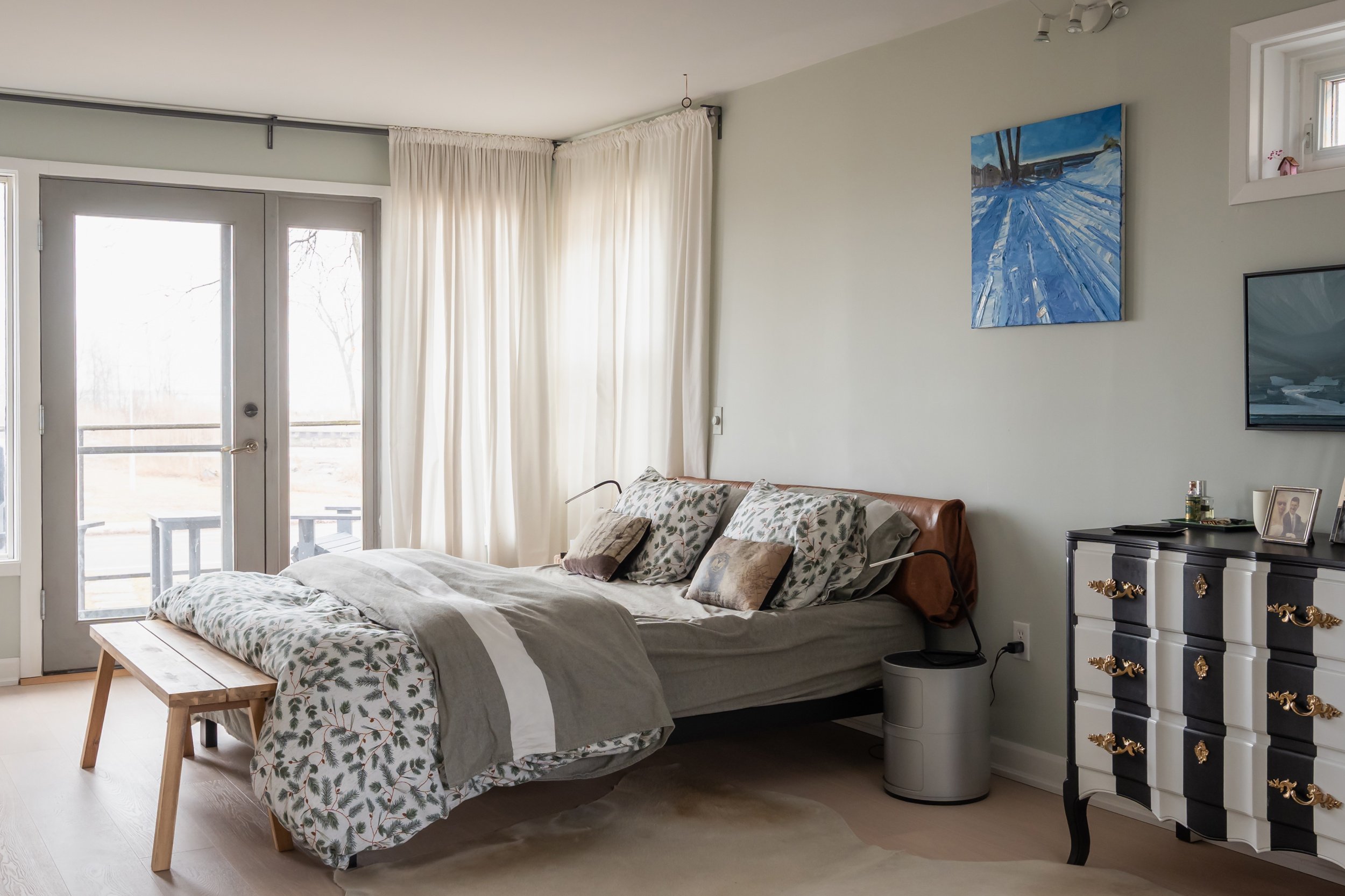 A bedroom with a bed, pillows, and a wooden bench at the foot of the bed near a large window with white curtains. There's a black and white dresser with gold handles, a TV, and framed photos. A landscape painting hangs on the wall.