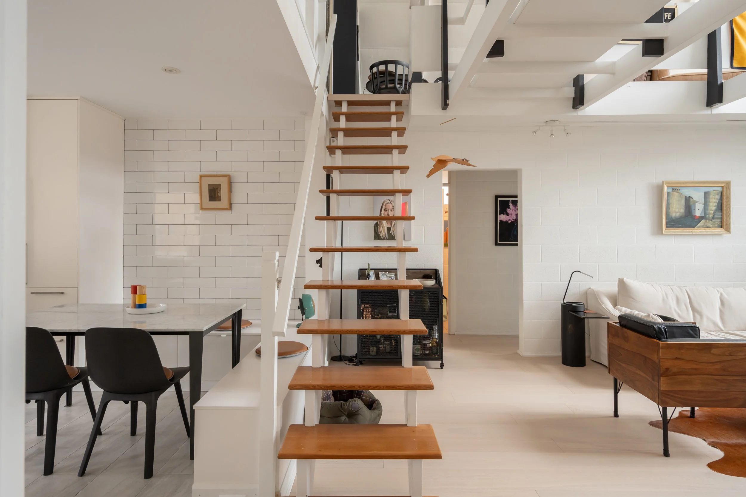 Living room with staircase, black dining chairs, marble table, white walls, wall art, and white sofa with wooden side table.
