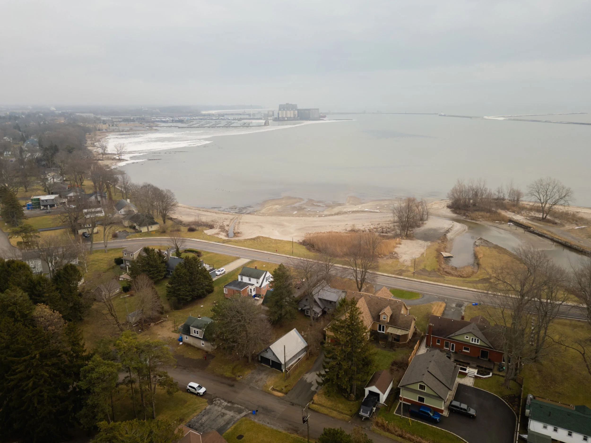 Aerial view of a neighborhood near a large body of water with some ice, trees, and houses, on a cloudy day.