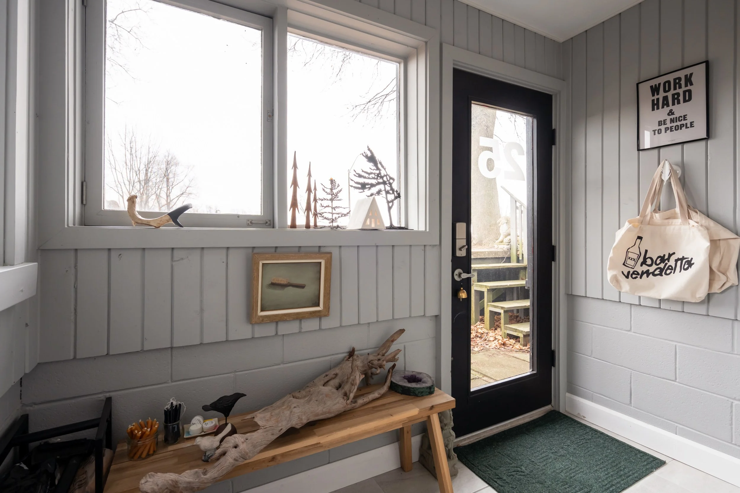 An entryway with gray paneled walls, a black door with large glass panel, and a window. Decor includes a framed picture of a spoon, small wooden trees and house outside, a large driftwood sculpture on a wooden bench, a bag hanging on the wall with th