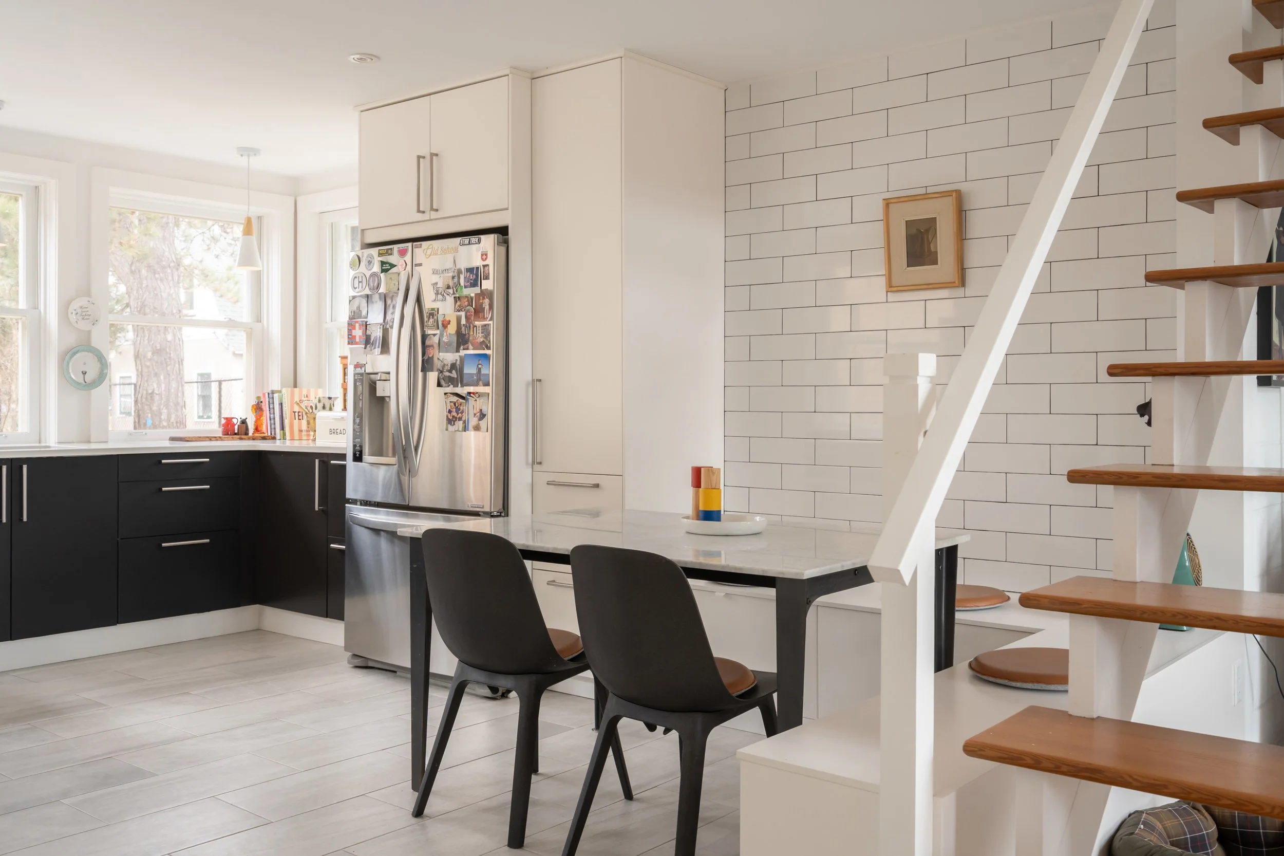 Modern kitchen with black lower cabinets, white upper cabinets, a stainless steel refrigerator covered in magnets, white subway tile backsplash, a dining table with black chairs, and wooden staircase with open risers.