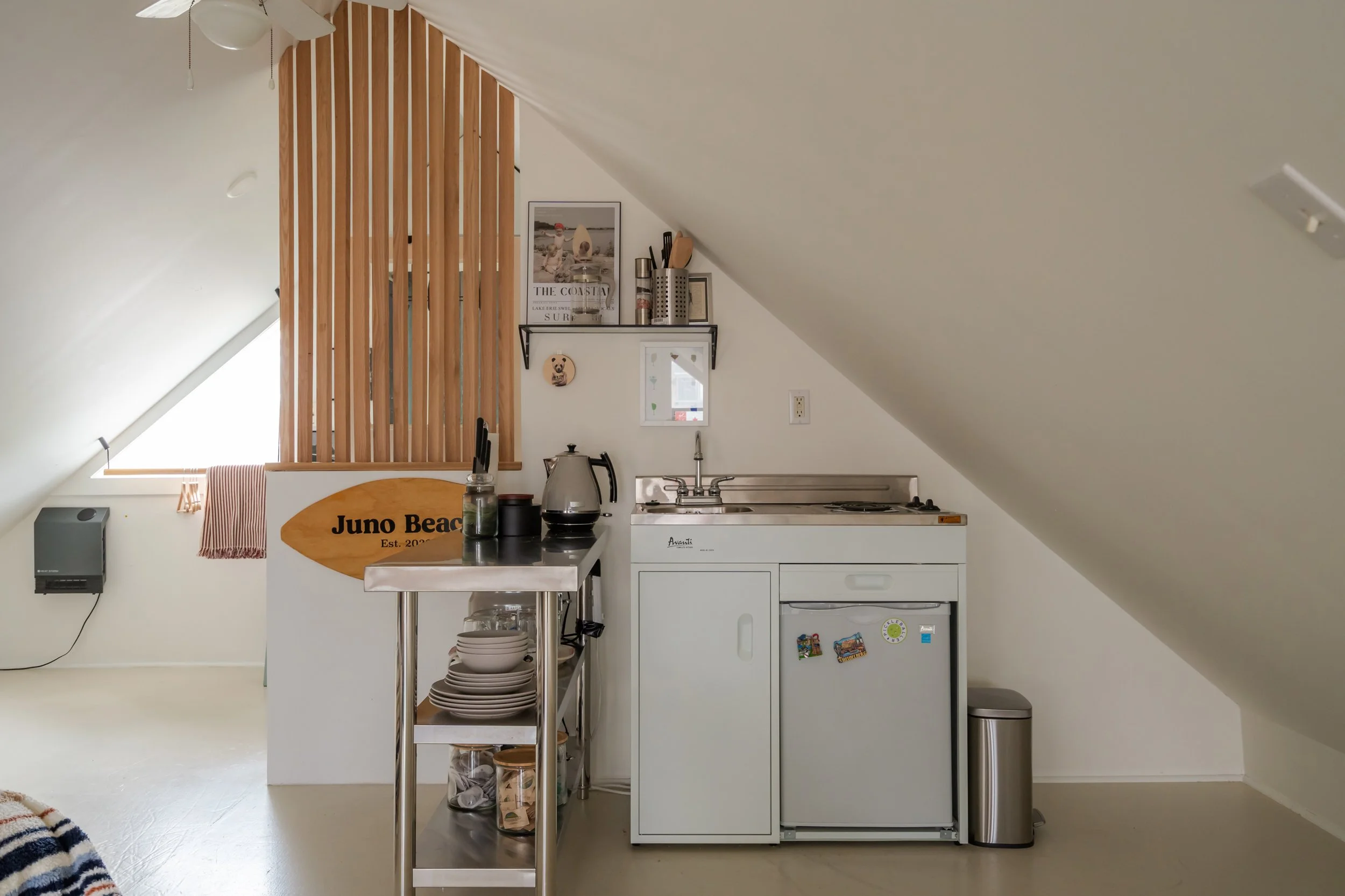 Small kitchen area in a room with a sloped ceiling, featuring a mini fridge, a small sink, and a countertop with a kettle and utensils. Behind, a sign reads "Juno Beach" and a wooden lattice separates the kitchen from a window with a pink towel hangi