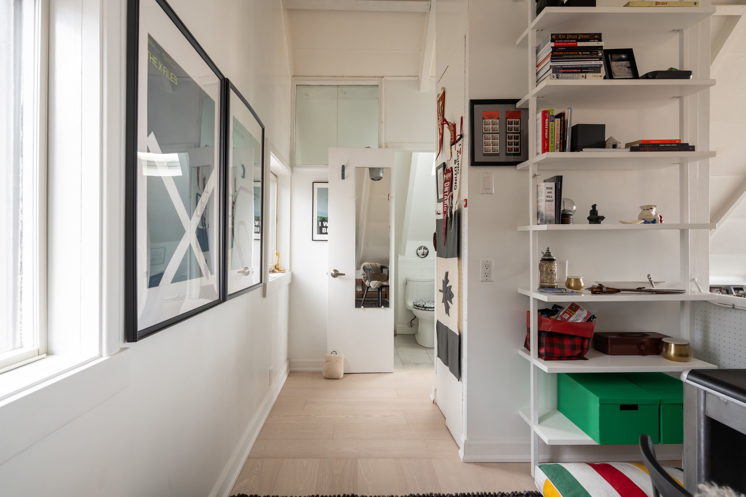 A hallway with artwork on the left wall, a door at the end, and a bathroom with a toilet visible through the open door. On the right, there is a white bookshelf filled with books, decorative items, and storage boxes.