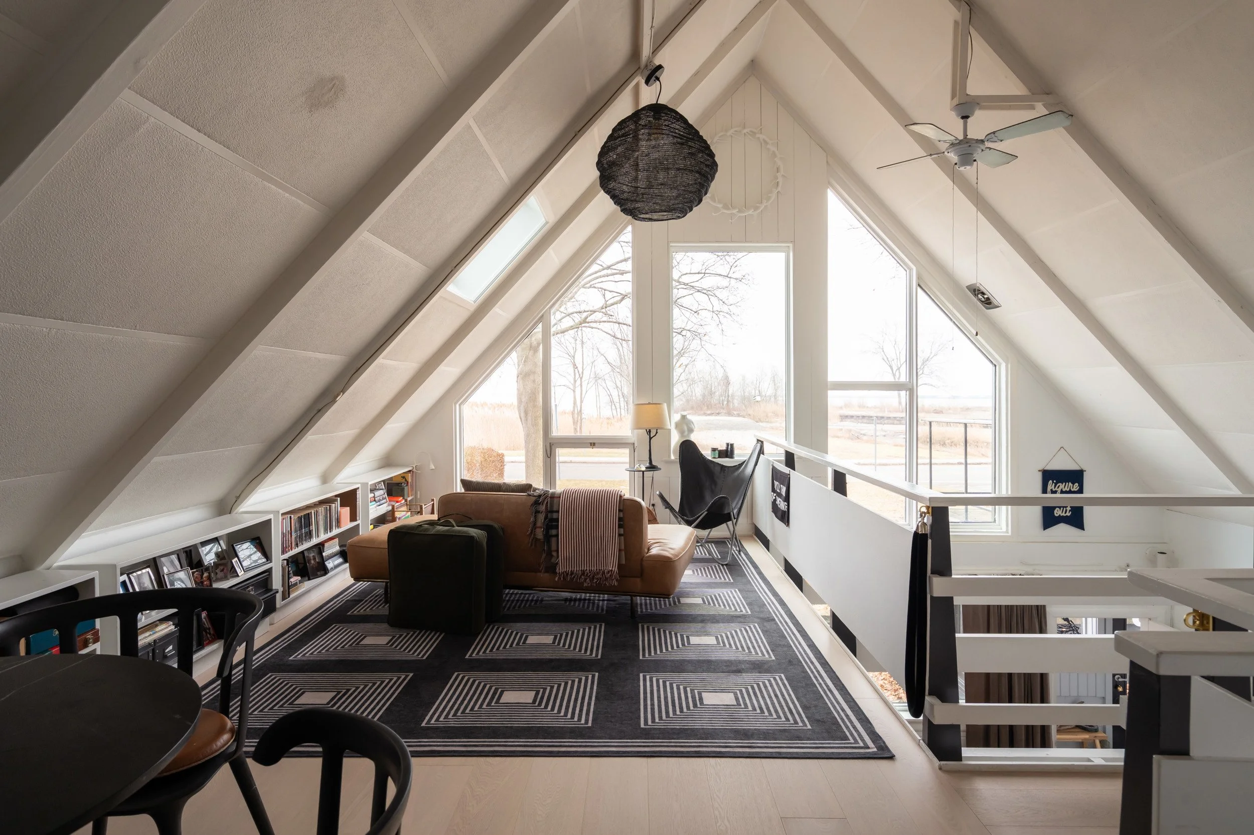 A cozy attic living space with angled white walls and large windows, featuring a brown sofa, black ottoman, bookshelf with photos and books, black and white geometric rug, black wingback chair, small table with a lamp, and a ceiling fan.