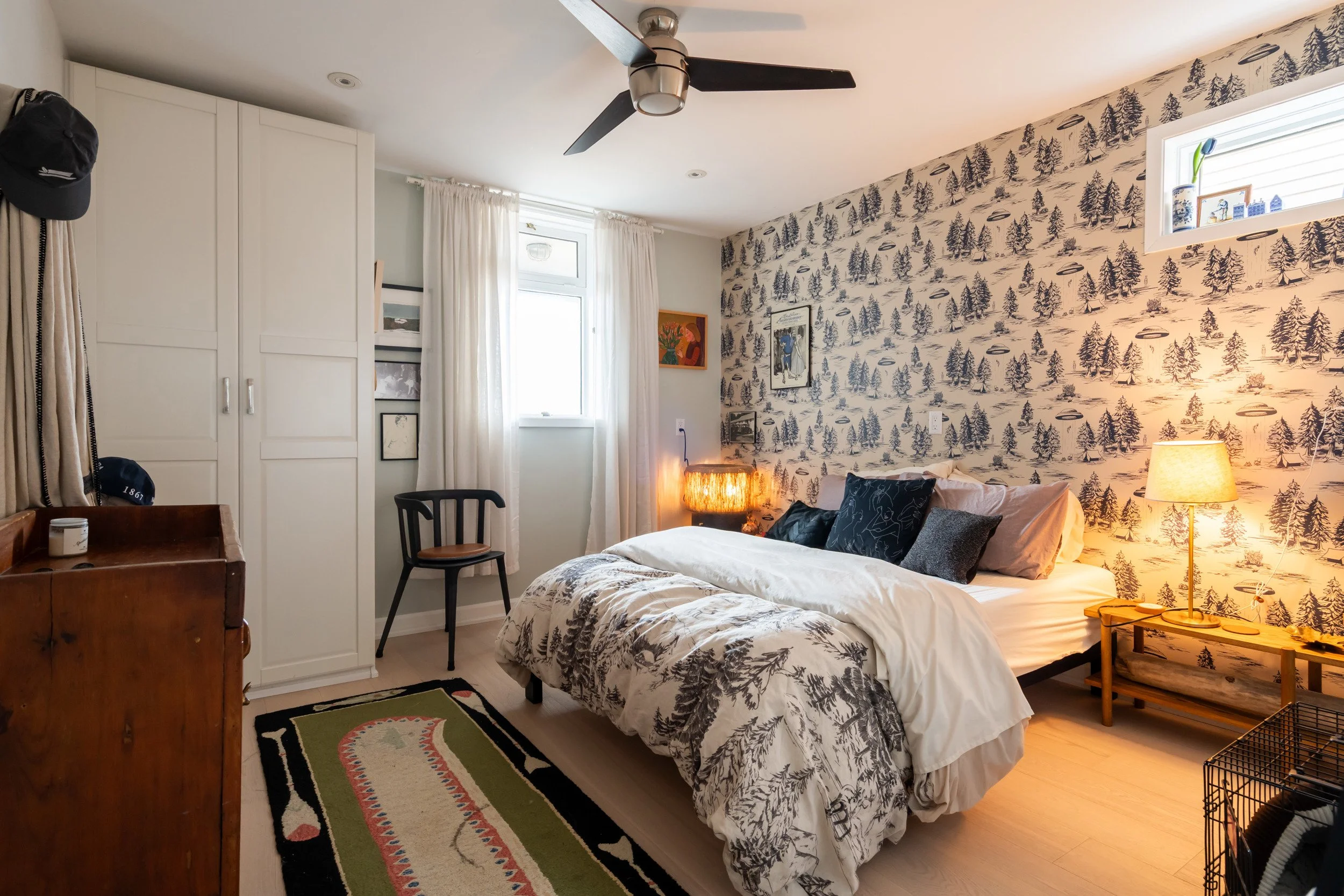A bedroom with a bed, black and gray pillows, and patterned bedding against a nature-themed wallpapered wall. There is a nightstand with a lamp, a small window, and a ceiling fan.