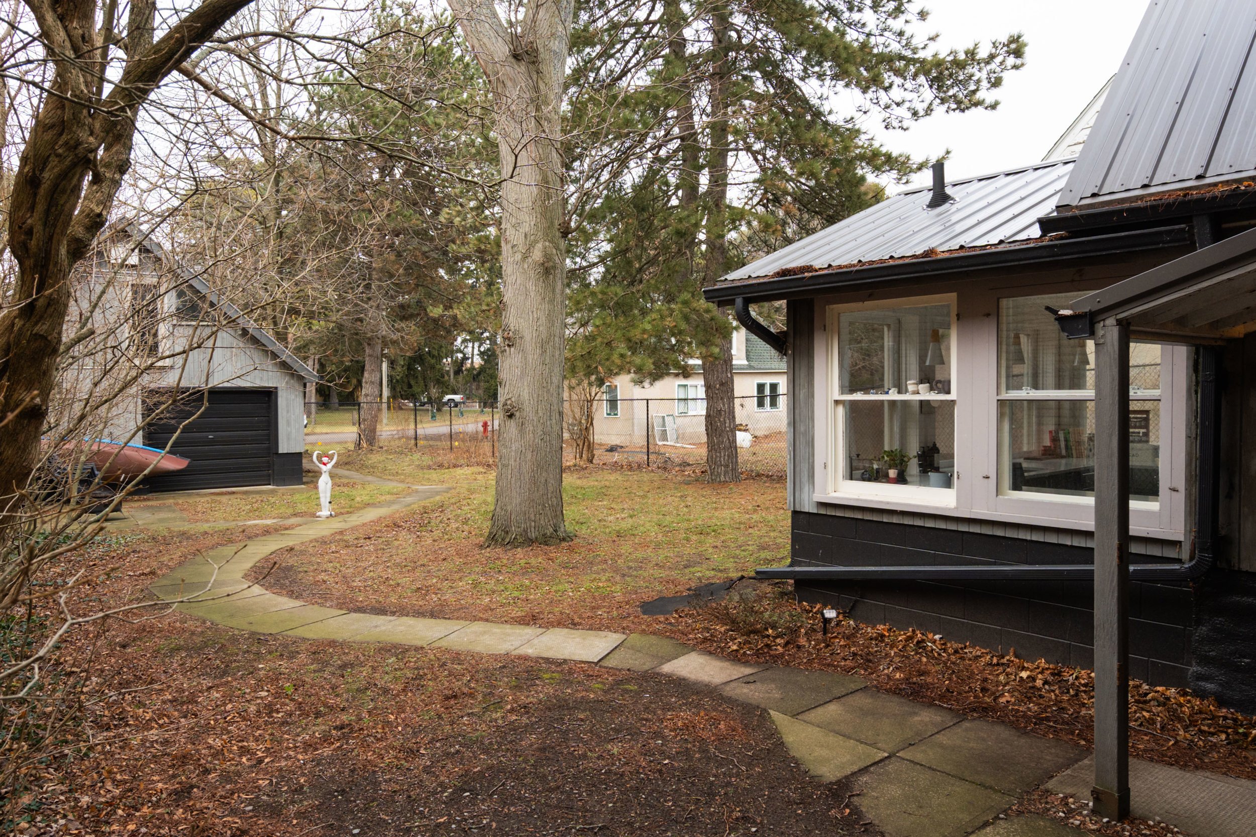 A backyard scene with a winding stone pathway, trees, a small black shed, and a house with large windows. There is a white sculpture of a dog near the pathway and a small boat on the ground by the shed during an overcast day.