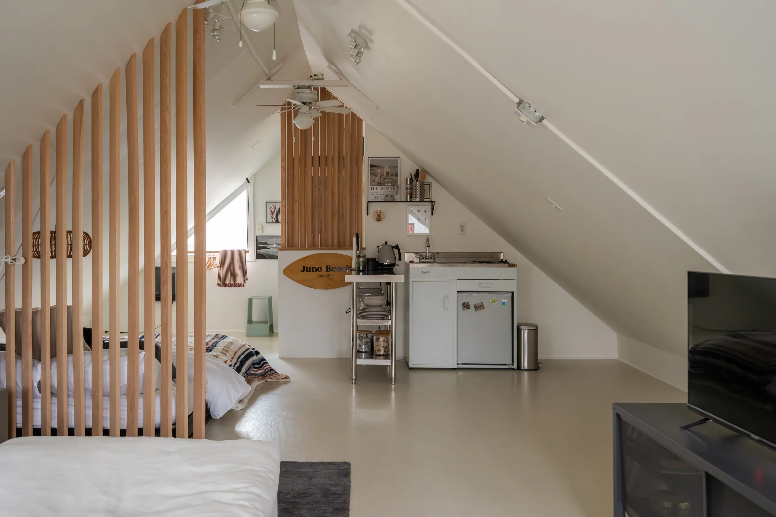 A small attic studio with a sloped ceiling, featuring a bed, a TV on a stand, a kitchenette with a mini-fridge, and a sign reading 'Juno Beach' on a wooden partition. There's minimal decor, track lighting, ceiling fans, and a window with a small curt