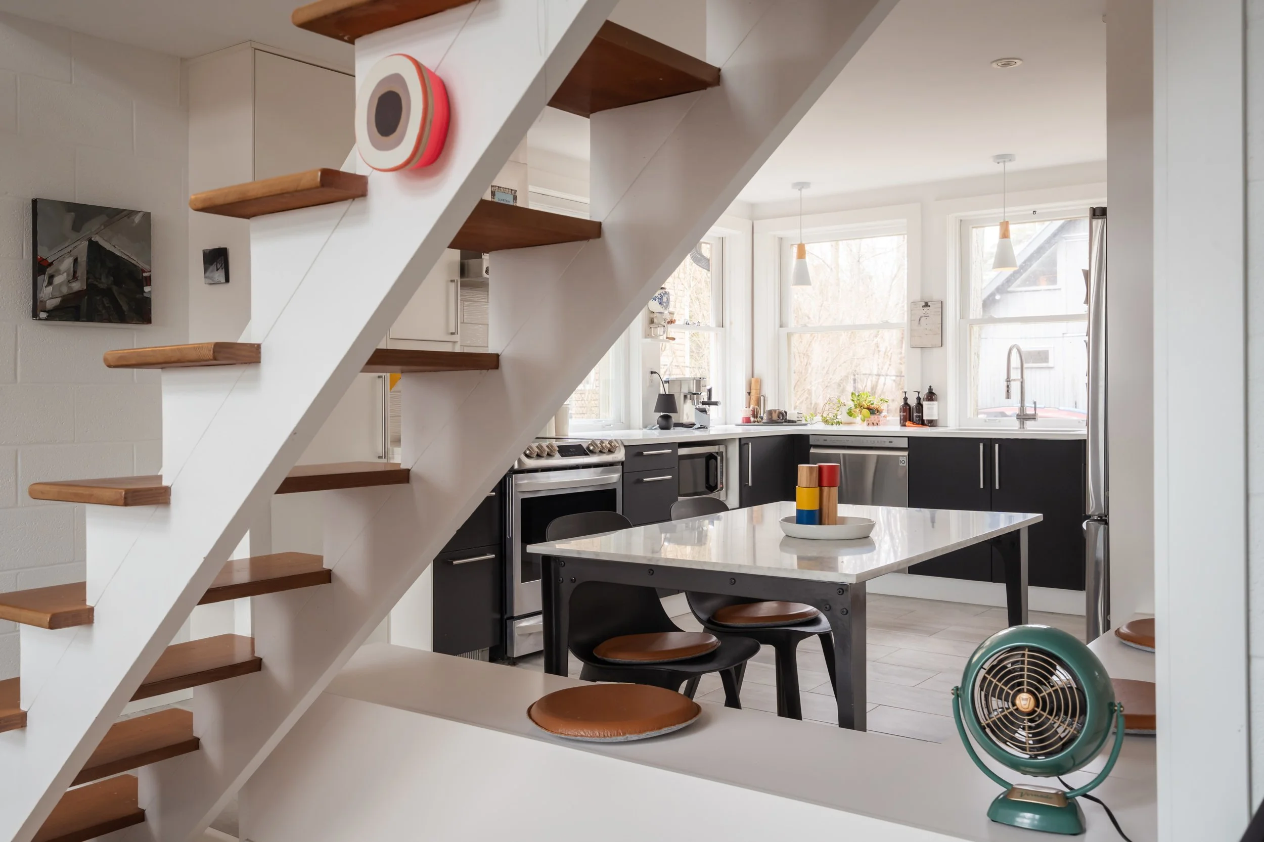 Modern kitchen with black cabinets, a white marble island, and large windows with a view of trees outside. A small round table with black chairs and brown cushions, decorative candles on the table, and a vintage green fan are visible. Open shelves, h
