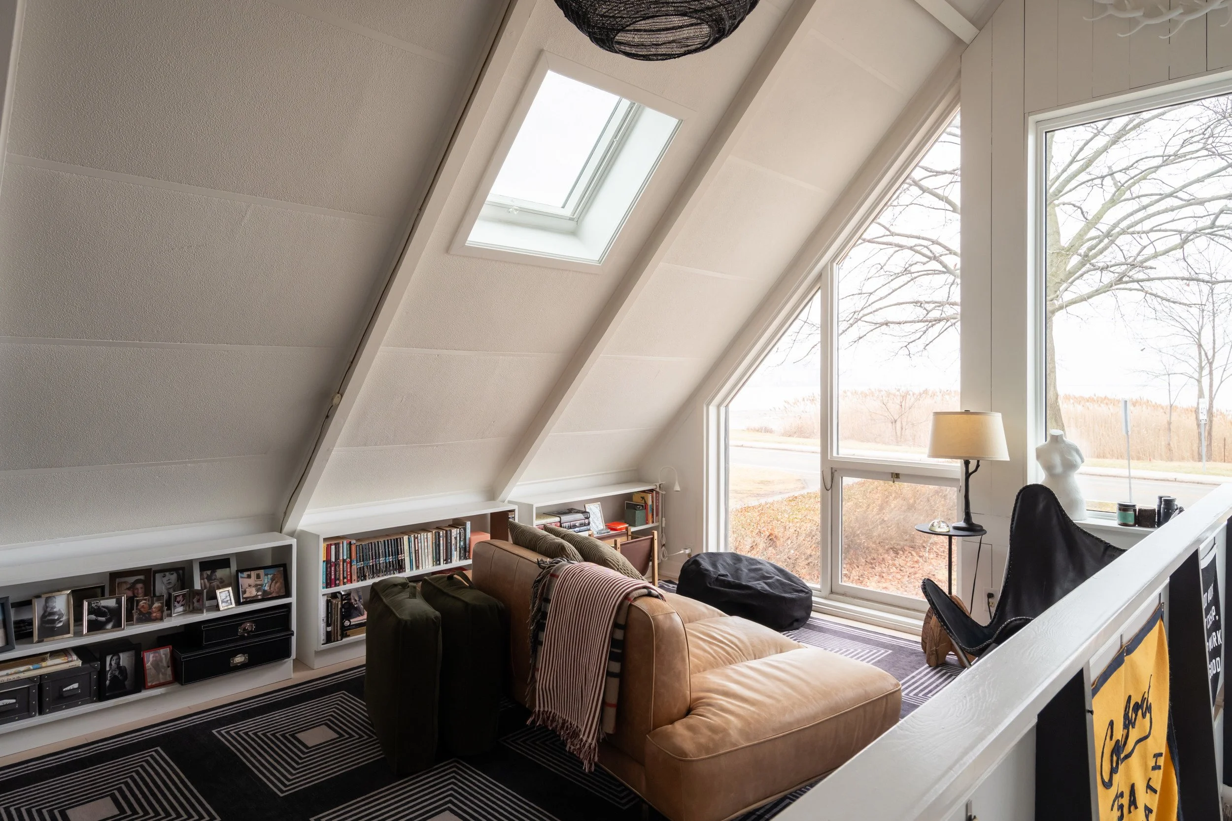 A cozy attic living room with a sloped ceiling, large windows, and a bookshelf.