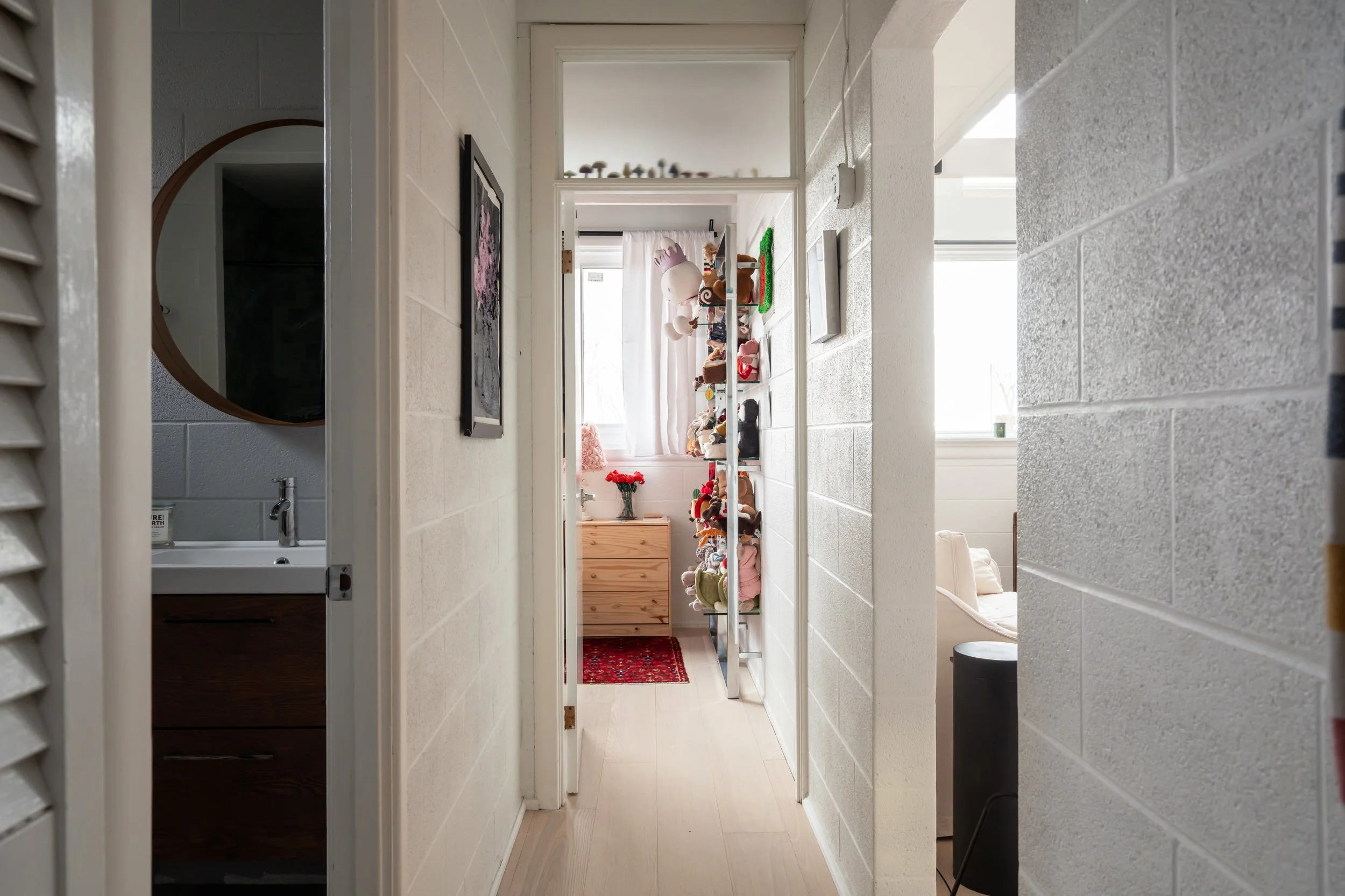 A narrow hallway leading to a room with a window, decorated with plush toys and stuffed animals on shelves and a small wooden dresser with a vase of red flowers, with natural light coming in through the window.