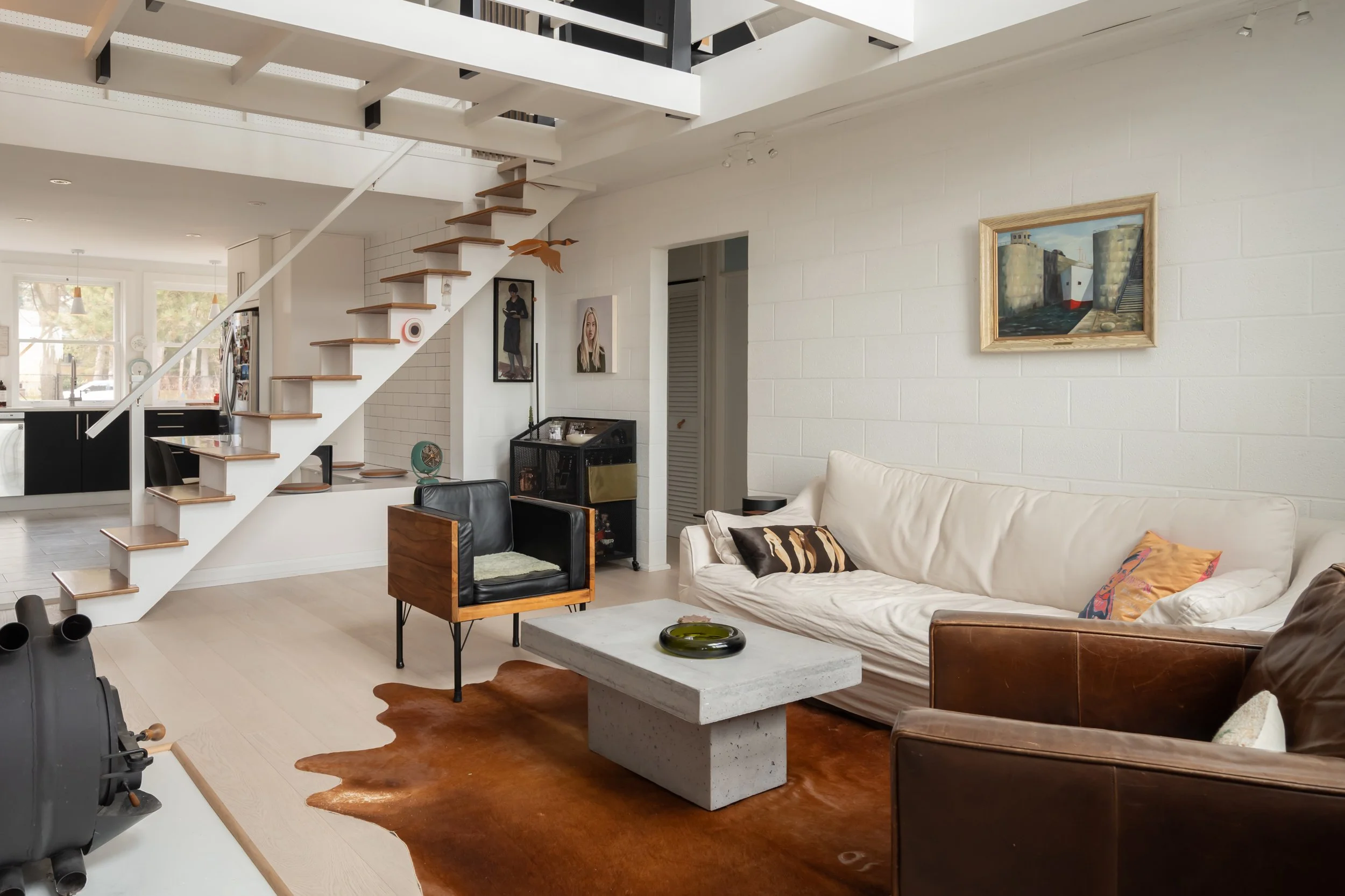 Living room with white brick wall, large white sofa with patterned pillows, brown leather armchair, concrete coffee table, cowhide rug, wall art, staircase with wooden steps, black kitchen in background.
