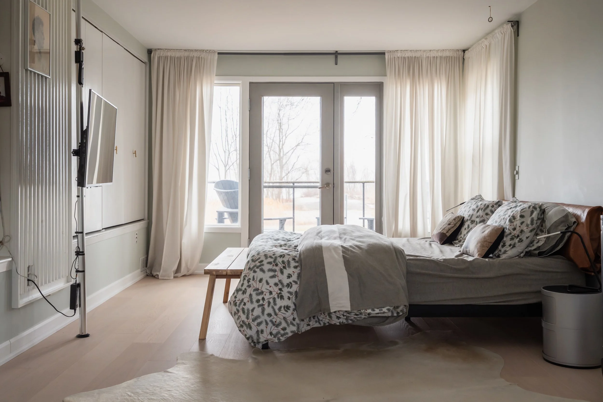 Sunlit bedroom with a bed, floral and neutral bedding, and cream curtains, leading to a balcony with outdoor seating, and a mounted television on the left.
