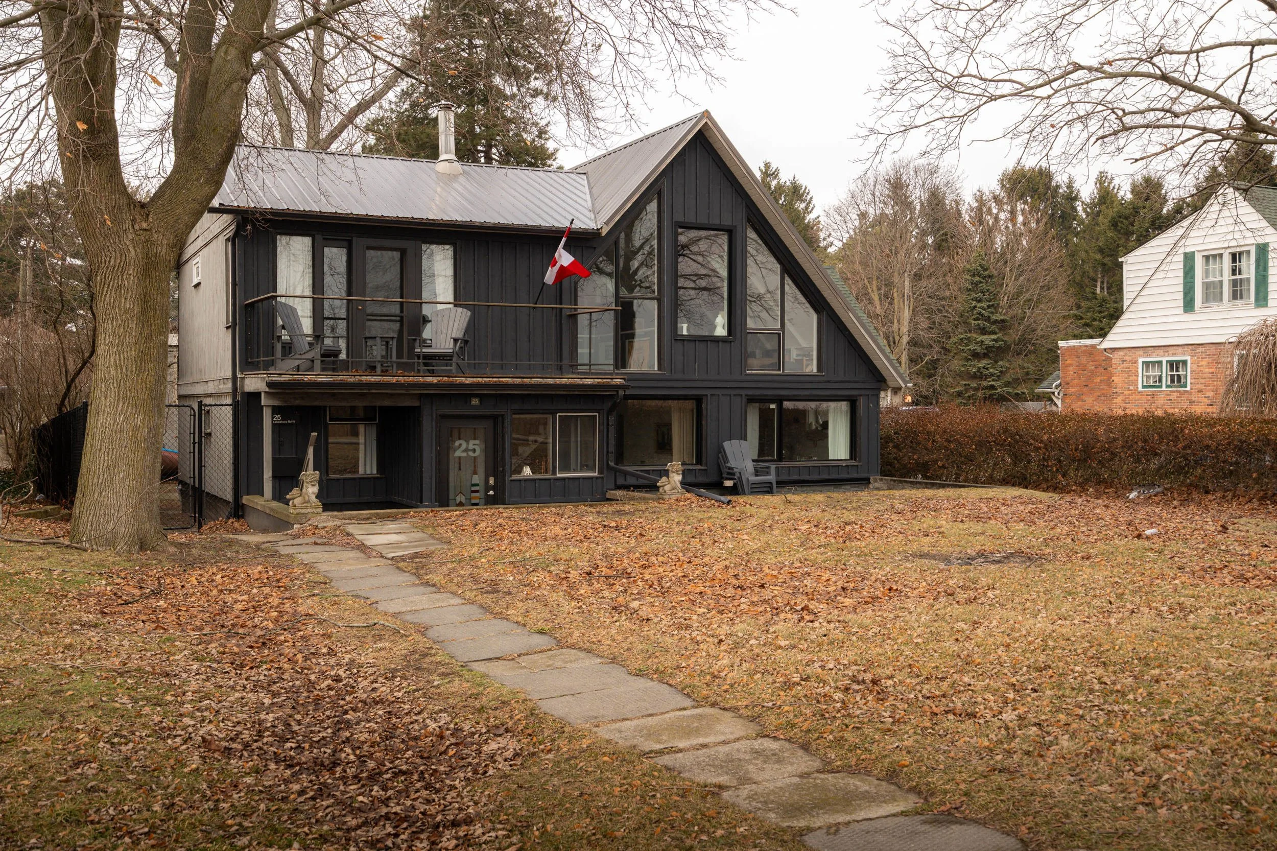 A two-story black house with a large triangular front window and a balcony, surrounded by fallen leaves and a few outdoor chairs, with a large tree to the left and neighboring houses in the background.