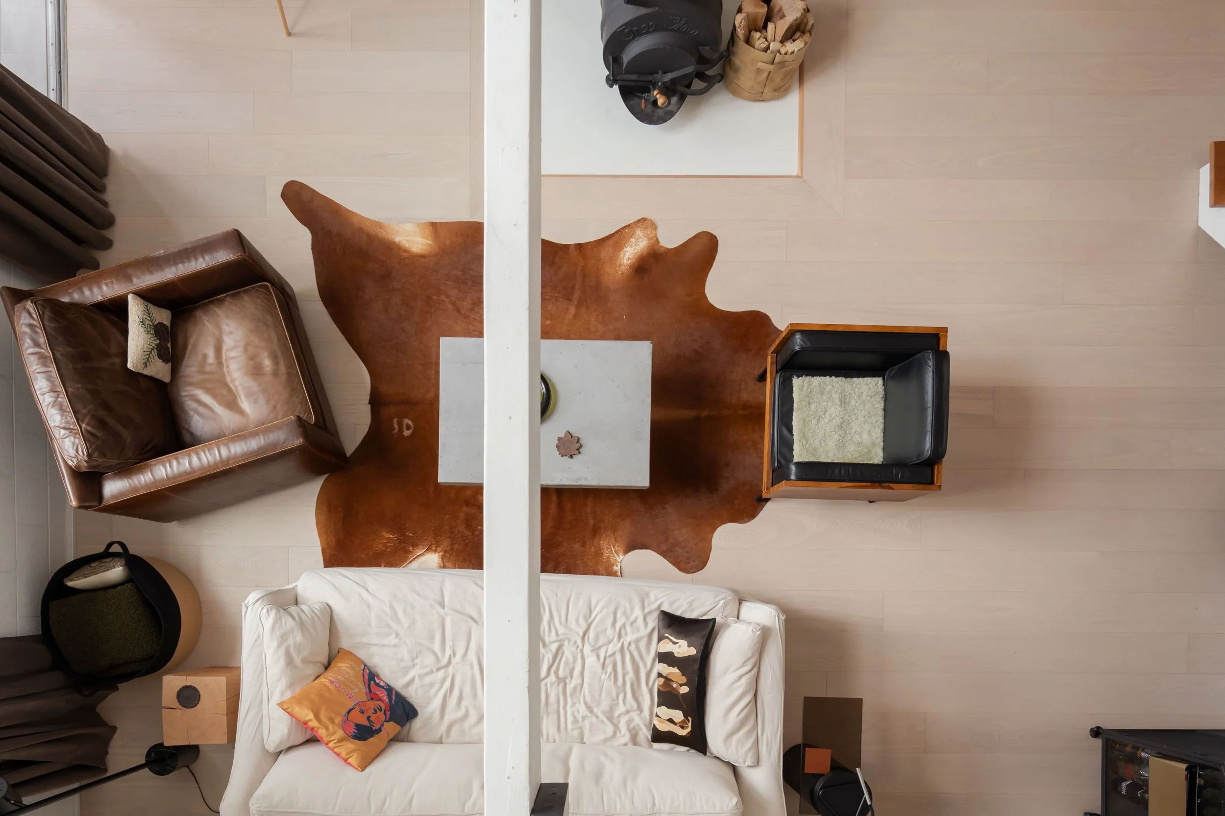 Top-down view of a living room with a leather armchair, a white sofa with pillows, a cowhide rug, a small coffee table, and various accessories, with a white beam dividing the image.