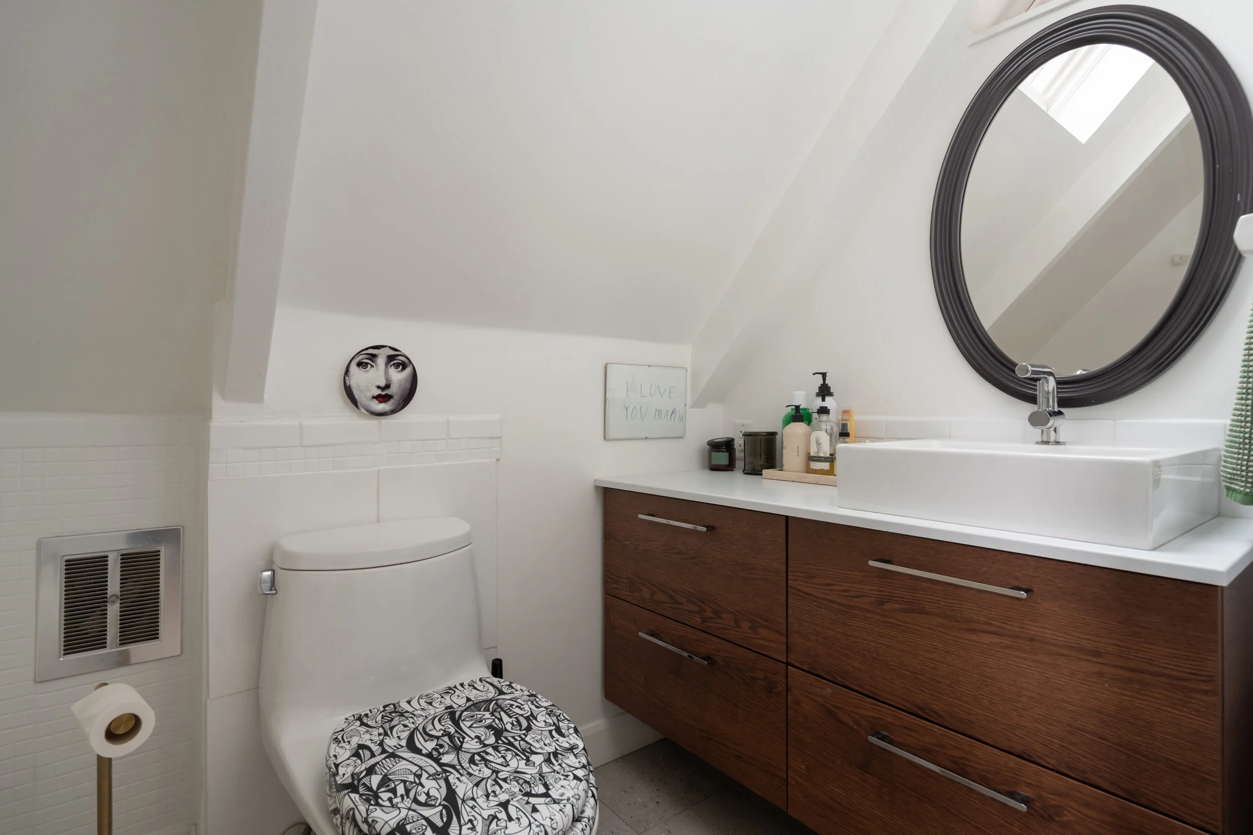 A modern bathroom with a white sloped ceiling, a white toilet with a black and white patterned seat cover, a wooden vanity with four drawers and a white countertop, a white vessel sink, a round black mirror, and various toiletries on the countertop. 