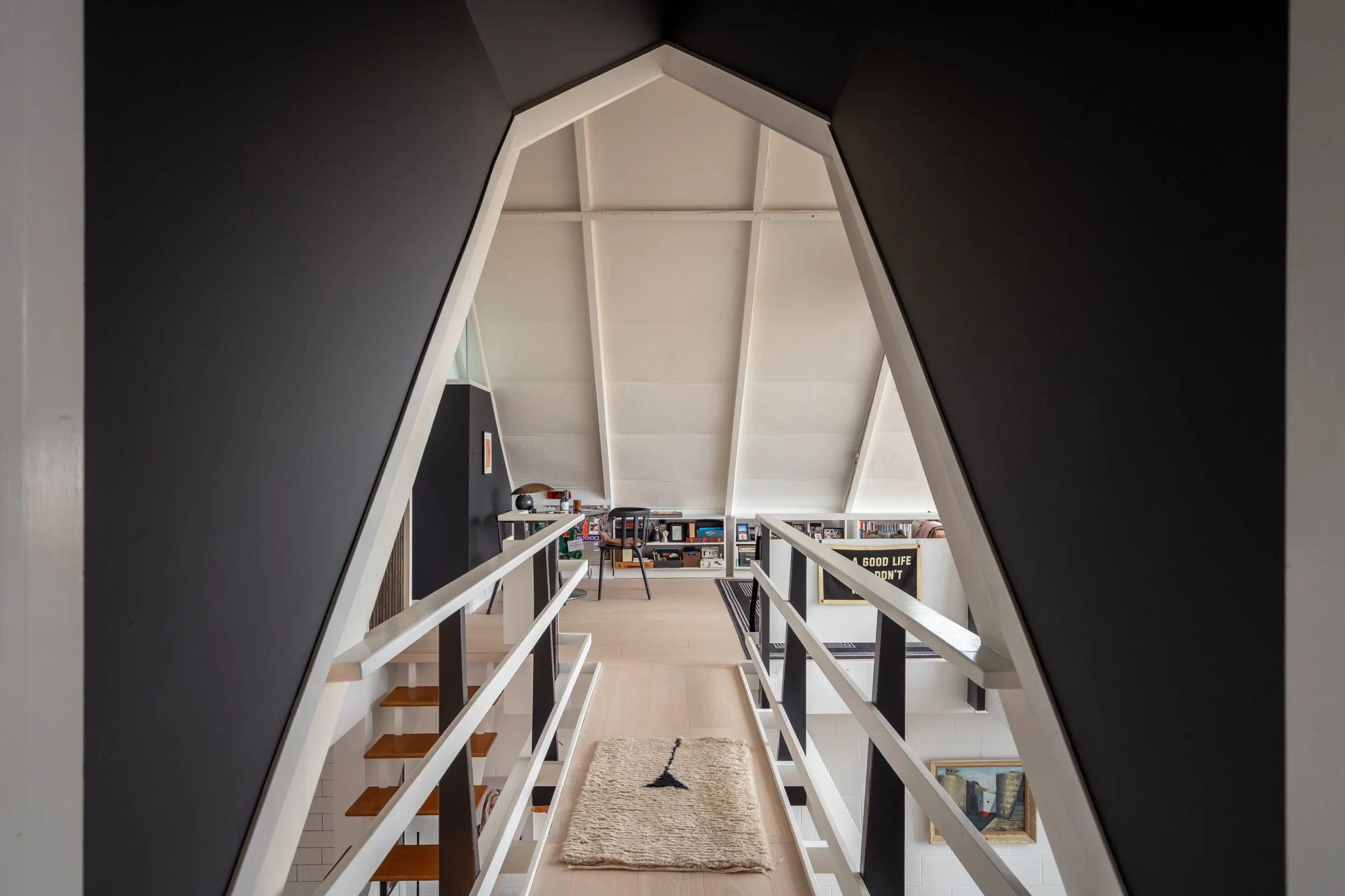 Interior view of a modern attic with a small mezzanine. The ceiling is sloped with white panels, and there is a black wall on the left. A beige rug with a black tree design is on the wooden floor, leading to a workspace area with shelves filled with 