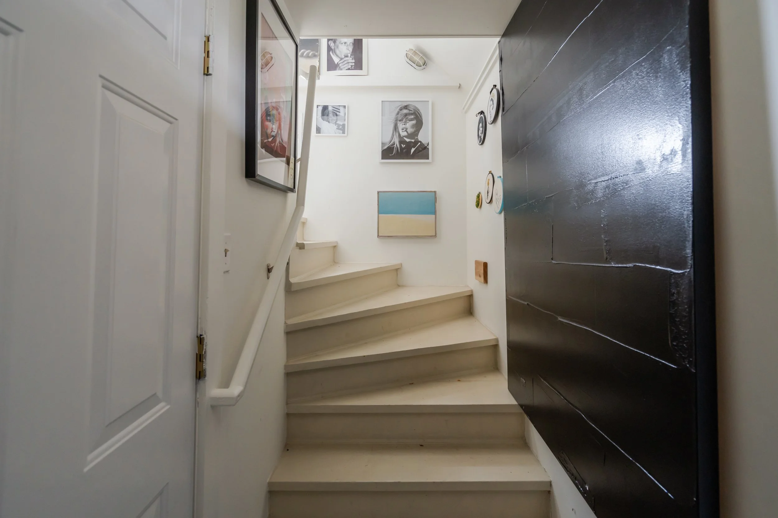 Interior view of a staircase with artwork and photographs on the walls, including two black and white portraits, and a painting of a beach scene. The stairs are beige, with a white wall to the left and a large black panel on the right.