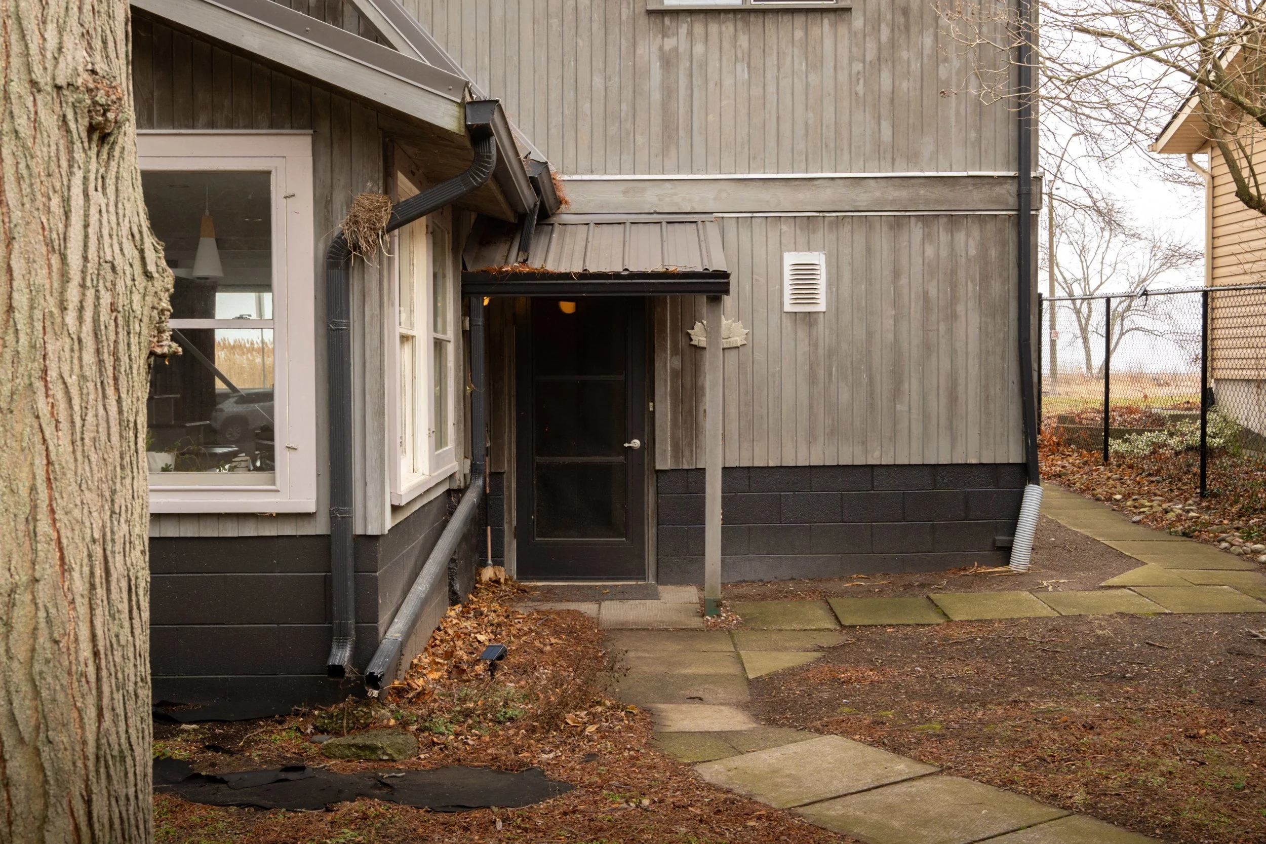 Backyard of a house with gray wooden siding, black foundation, and black drainage pipes, with fallen leaves and a concrete pathway.