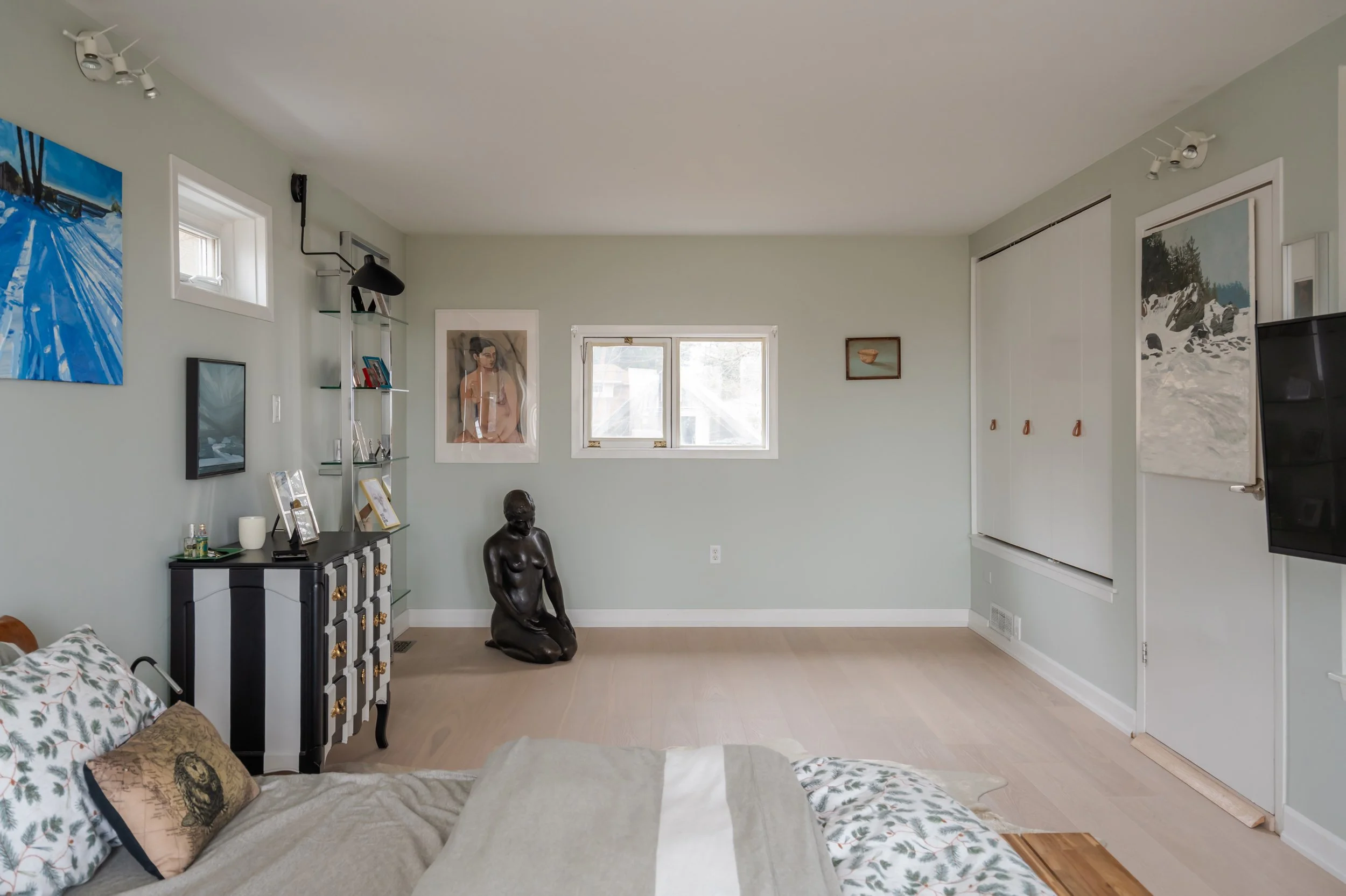 A living room with a bed in the foreground, decorated with floral pillows. There are artworks on the wall, a black and white dresser, a black sculpture of a kneeling woman, and a wall-mounted TV. The room has light green walls, wooden flooring, and a