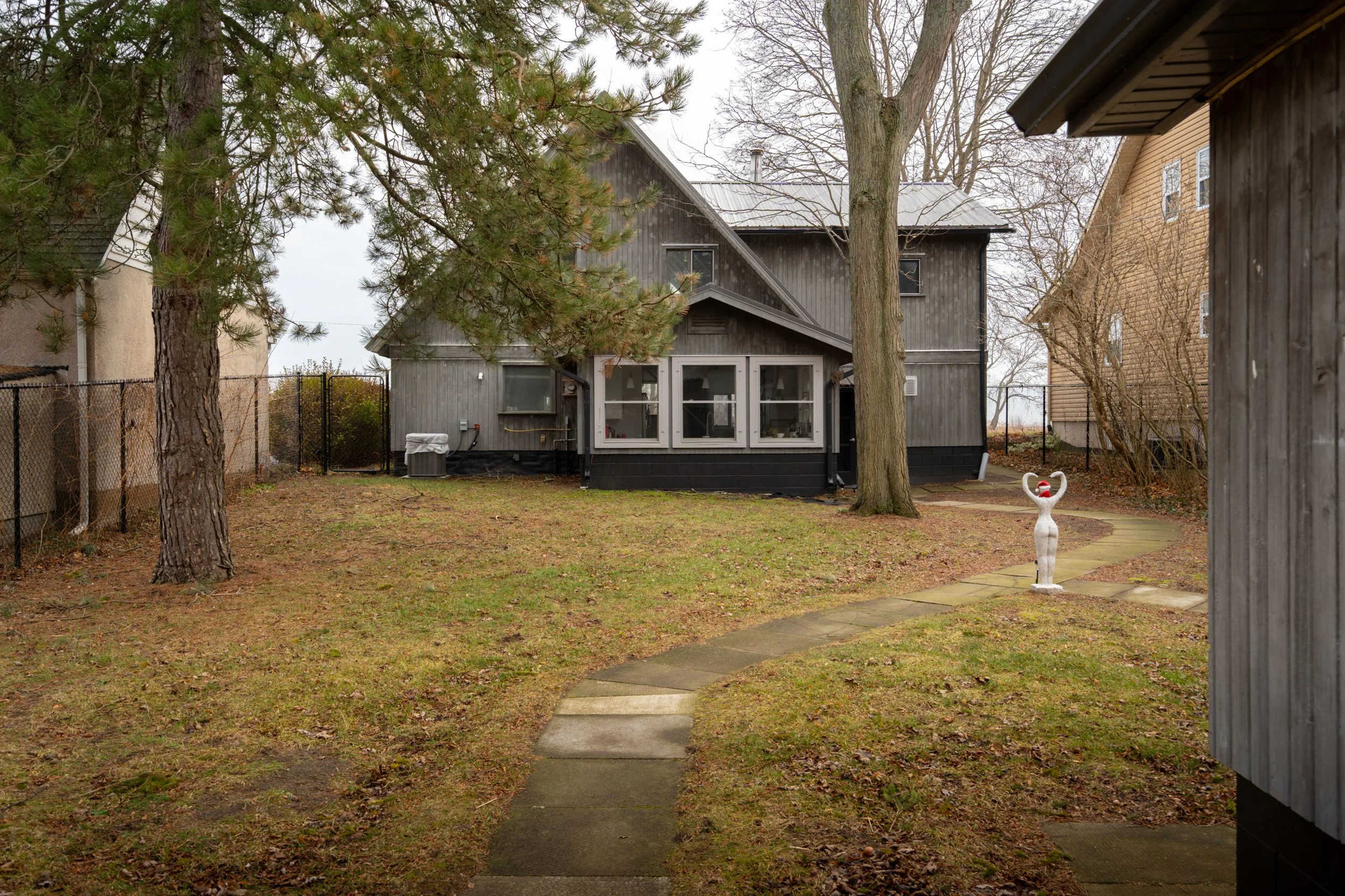 Backyard with a concrete pathway, trees, a wooden house, and a garden statue of a woman with arms raised and a heart-shaped face.