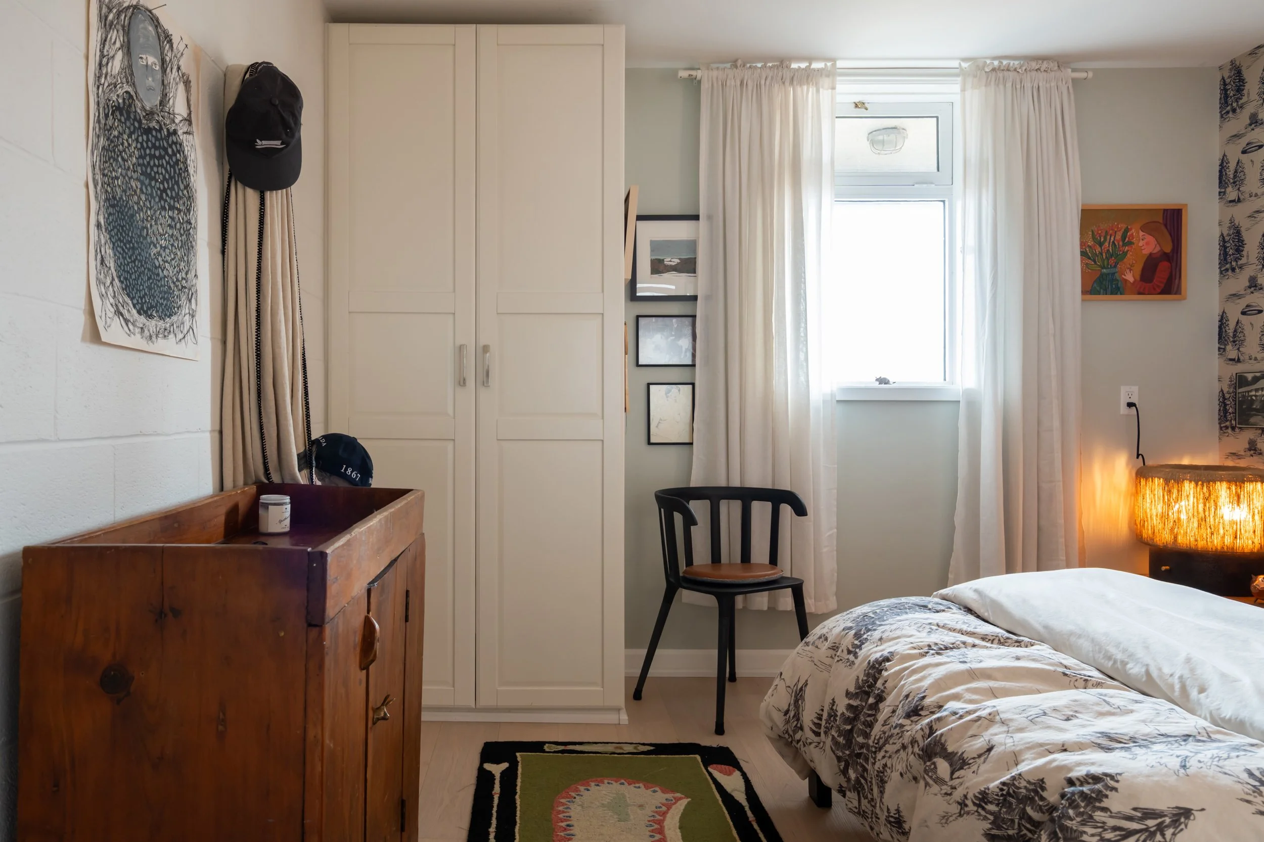 A cozy bedroom with white walls and light-colored wooden floor. There is a bed with a patterned black and white duvet on the right side. Next to the bed is a wooden nightstand with a warm, glowing lamp. On the left, a black chair with a round wooden 