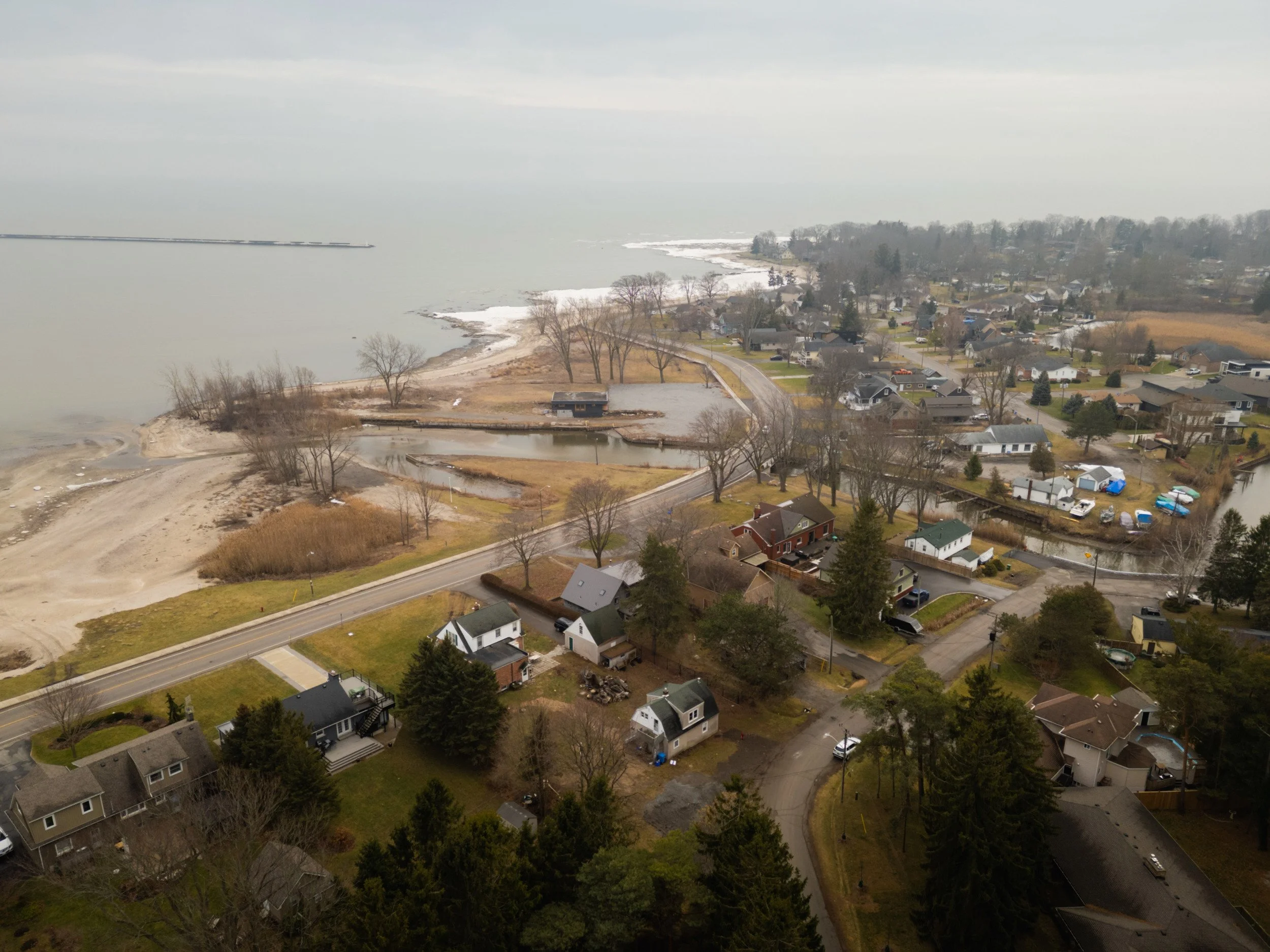 Aerial view of a coastal residential neighborhood with houses, trees, and roads, near a body of water with shoreline, small beaches, and a marina or pier in the background.