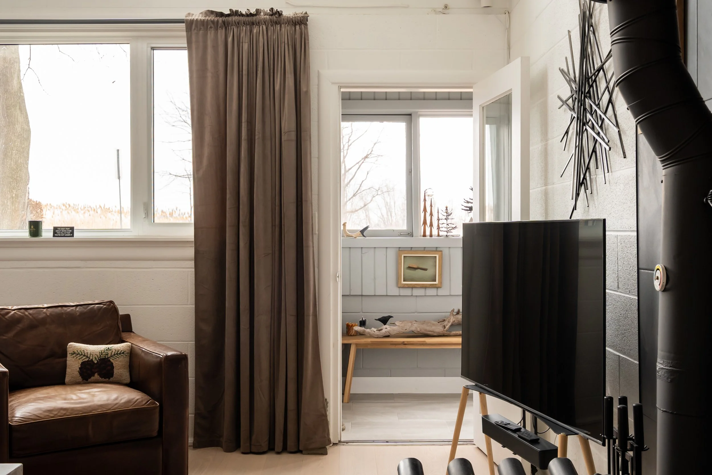 A living room with a brown leather sofa, a window with beige curtains, and a TV on a wooden stand. There is a small enclosed porch with a window, decorated with wooden art pieces and a framed picture on the wall.