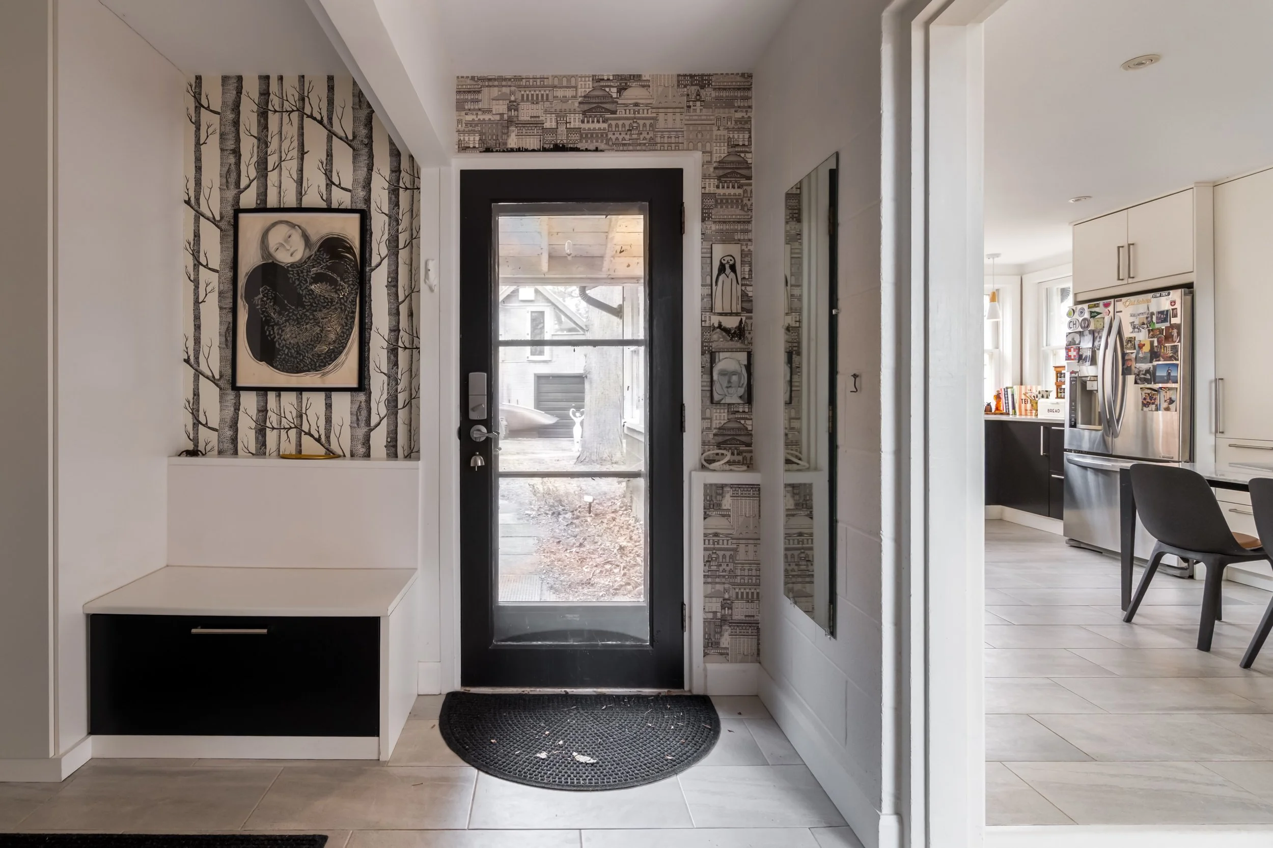 Entryway with a black door, framed artwork, patterned wallpaper, and a mirror. leading into a kitchen with a refrigerator and dining area.