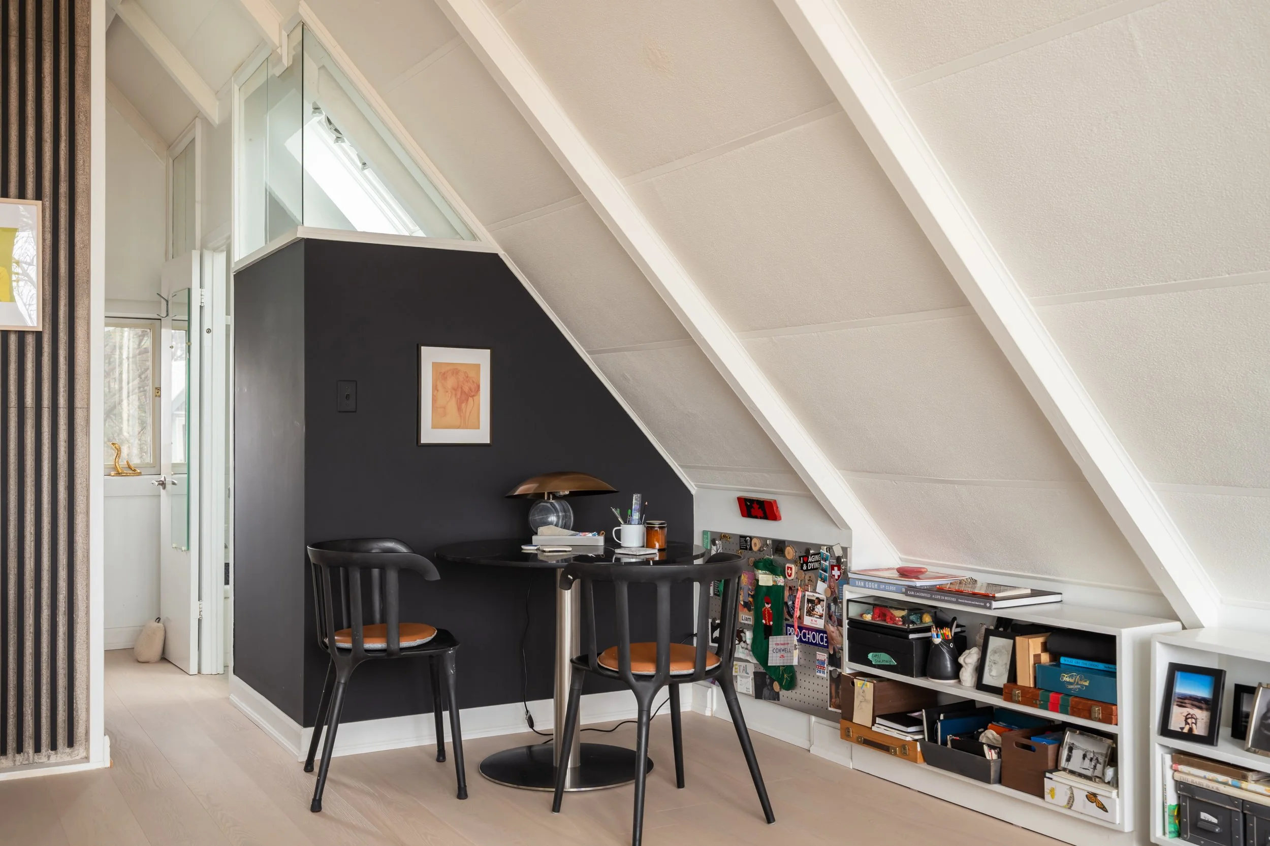 A cozy attic nook with a black-painted wall, a small black round table, and two black chairs with orange cushions. There's a small lamp, office supplies, and personal photos and decorations on a white built-in shelf along the sloped ceiling. A framed