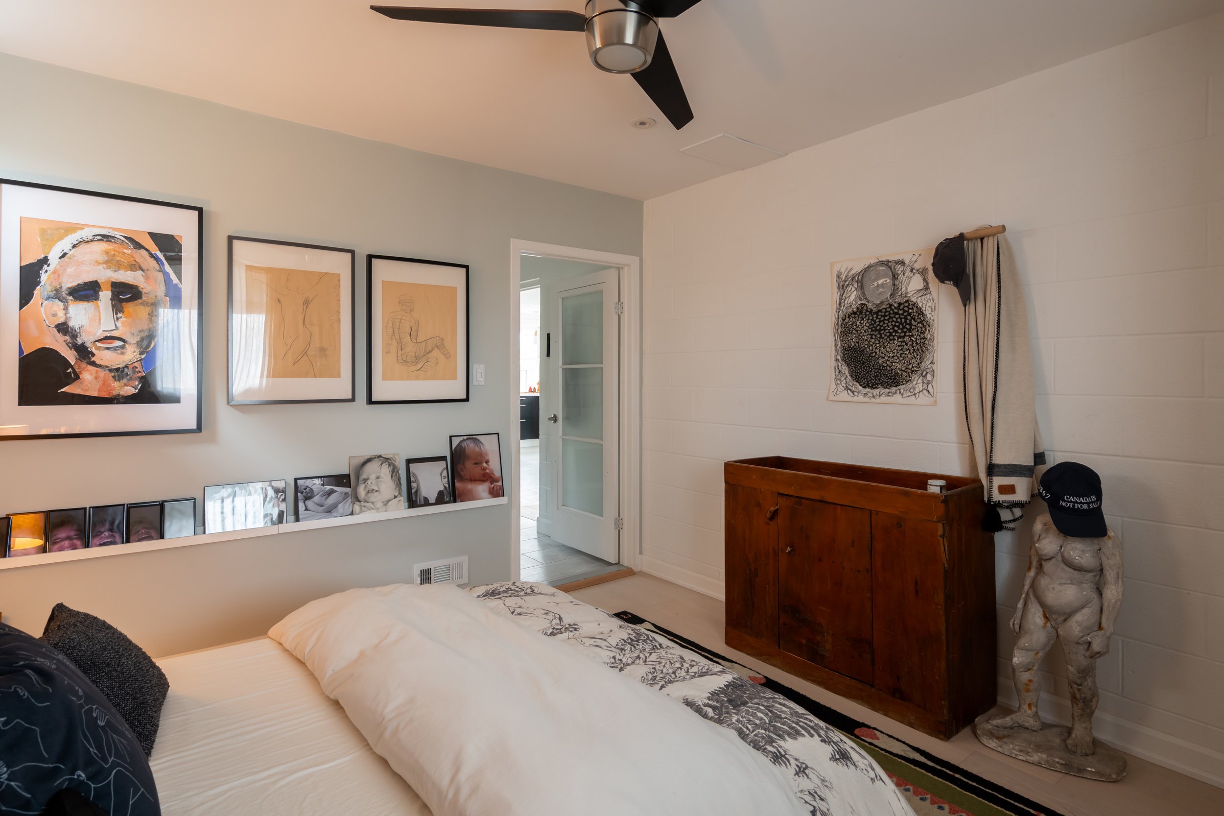 A bedroom with a bed, artwork on the walls, framed photos, a wooden cabinet, and a sculpture of a chubby figure wearing a baseball cap and holding a cap. The room has a ceiling fan and a door leading to another room.