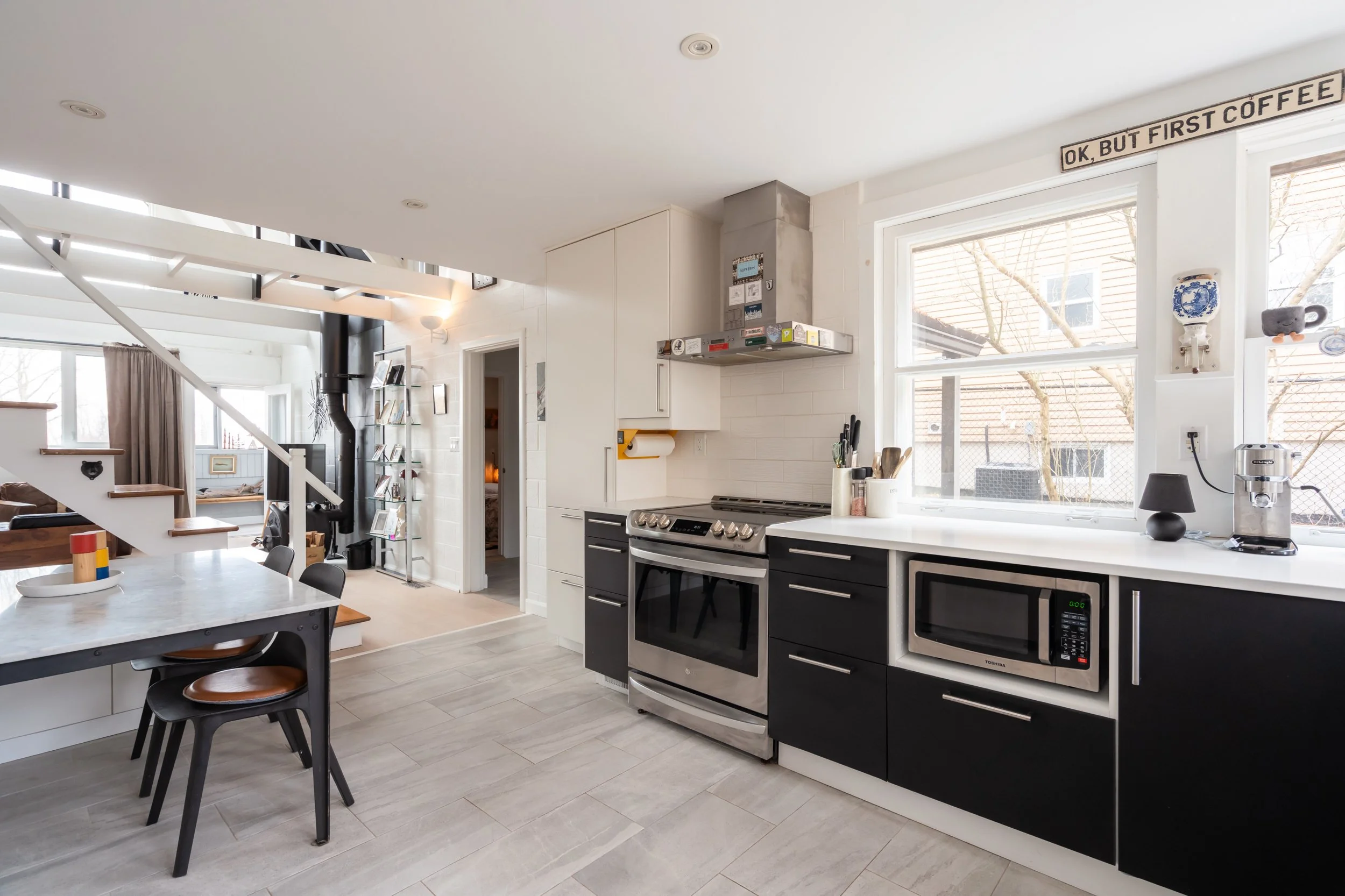 Modern kitchen with white and black cabinets, stainless steel oven, microwave, and coffee maker, with large windows showing trees outside.