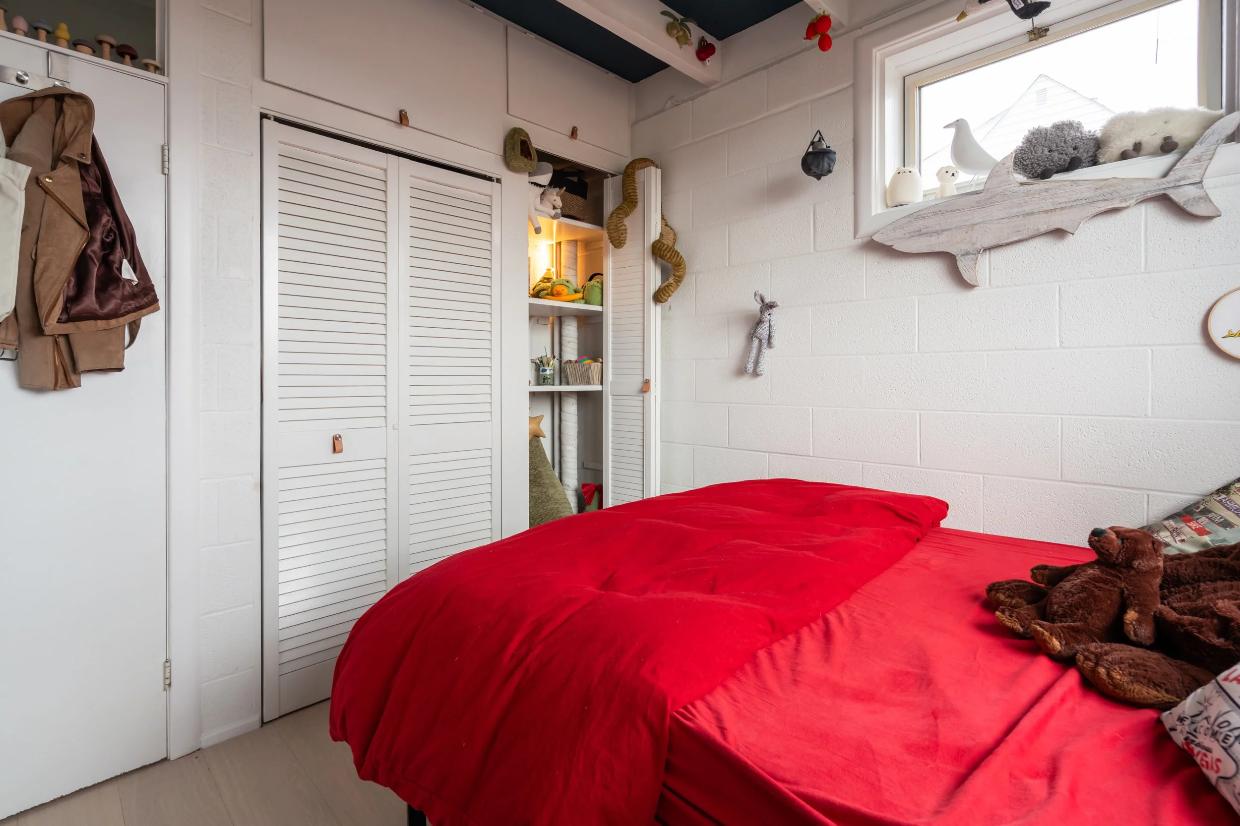 A cozy bedroom with a red bedspread, plush teddy bears on the bed, a window with decorative animals and felted figures, a white wall with hanging stuffed or decorative objects, and a closet with white louvered doors.
