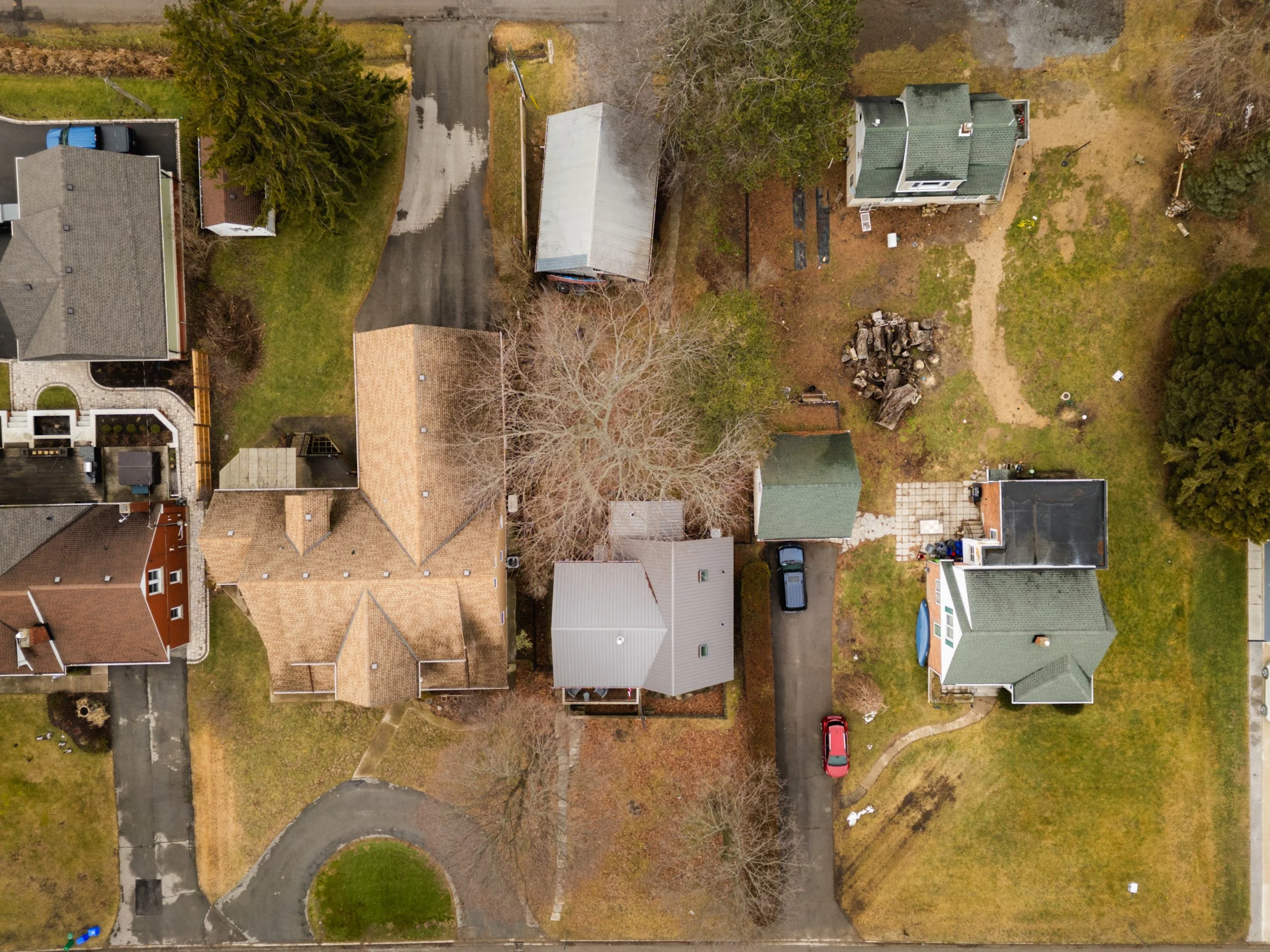 An aerial view of residential houses, yards, and driveways in a neighborhood during late fall or early winter, with some trees without leaves and patches of grass.