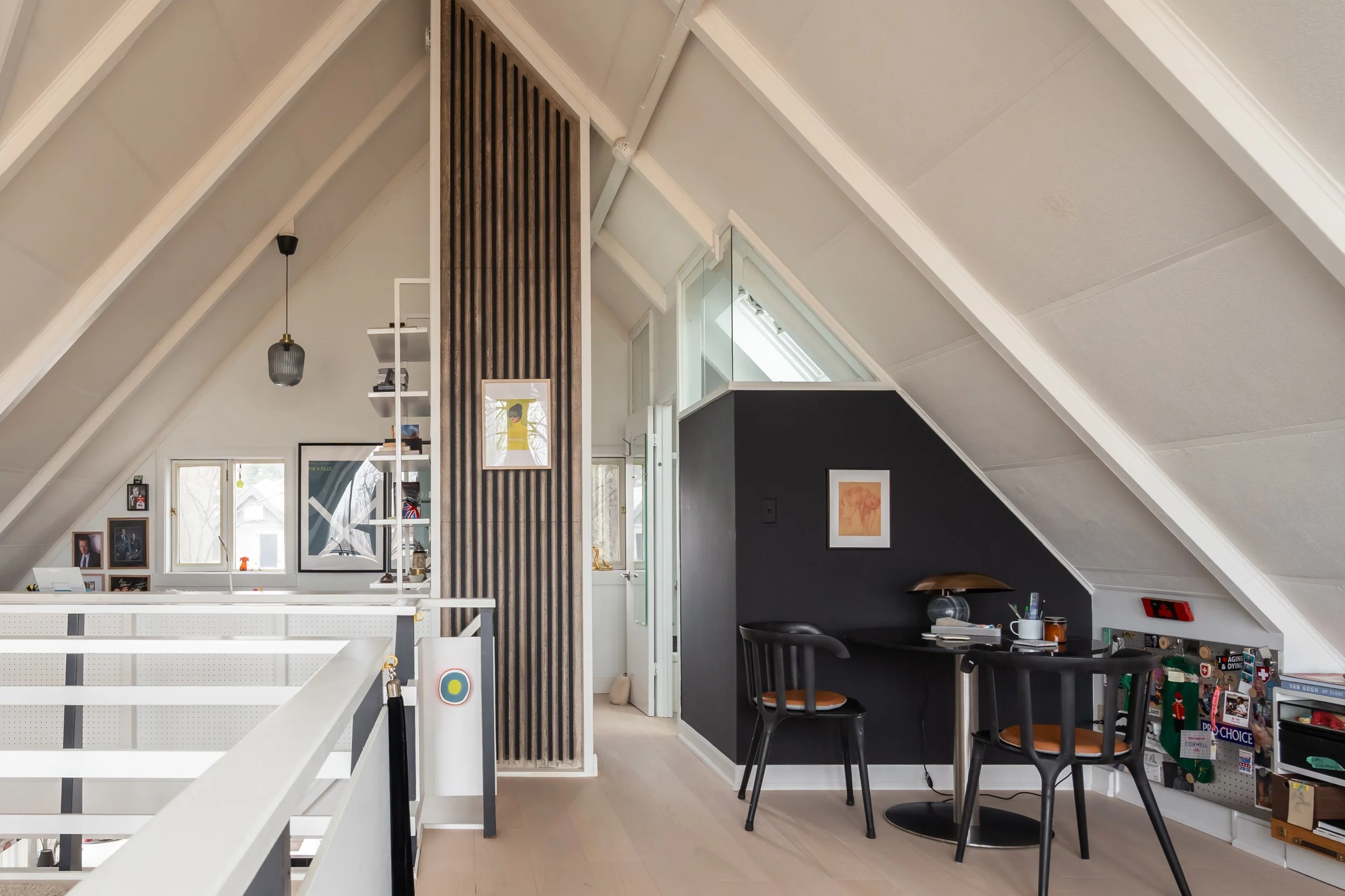 Interior view of an attic with sloped white ceiling, black accent wall, small desk with two black chairs, a table lamp, artwork on the wall, and shelves with books and decorations.