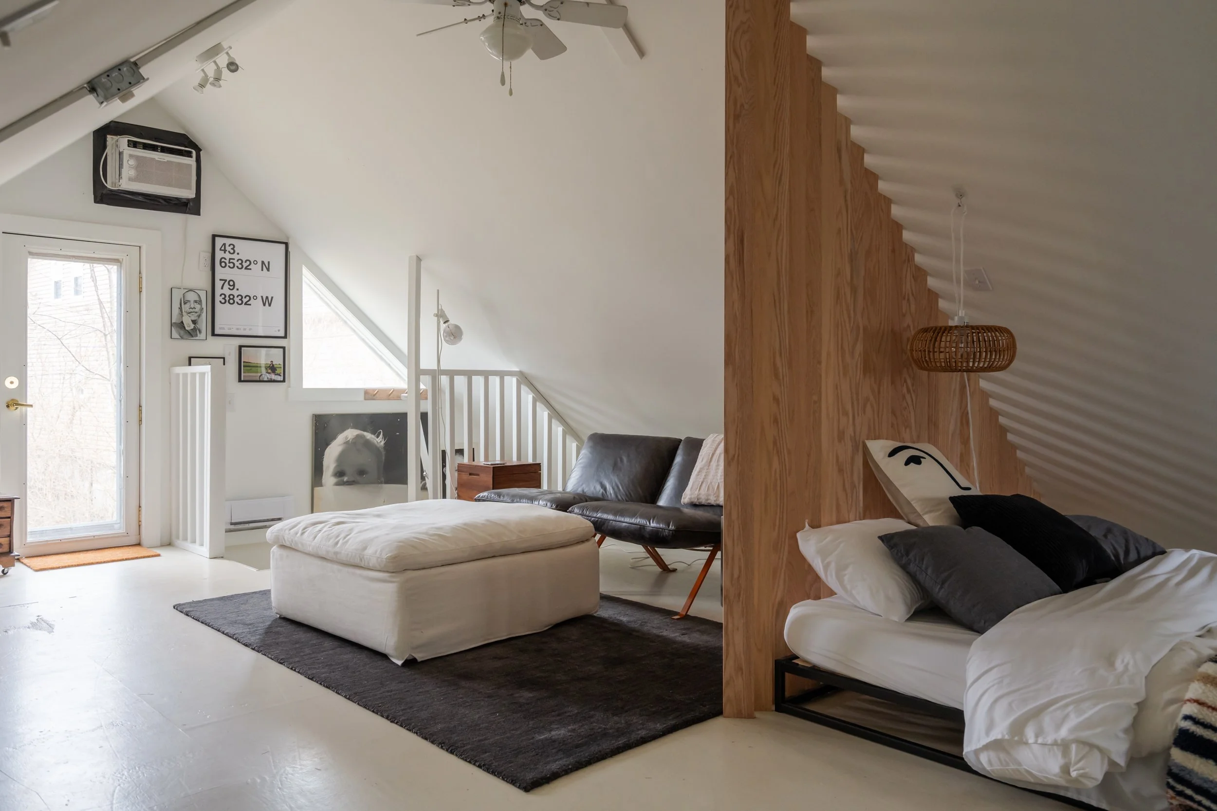 A modern attic apartment with sloped ceilings painted white. It features a black leather sofa, a white ottoman on a dark rug, and a bed with white and black bedding. There are wooden accents, including a wooden half-wall and small wooden side tables.