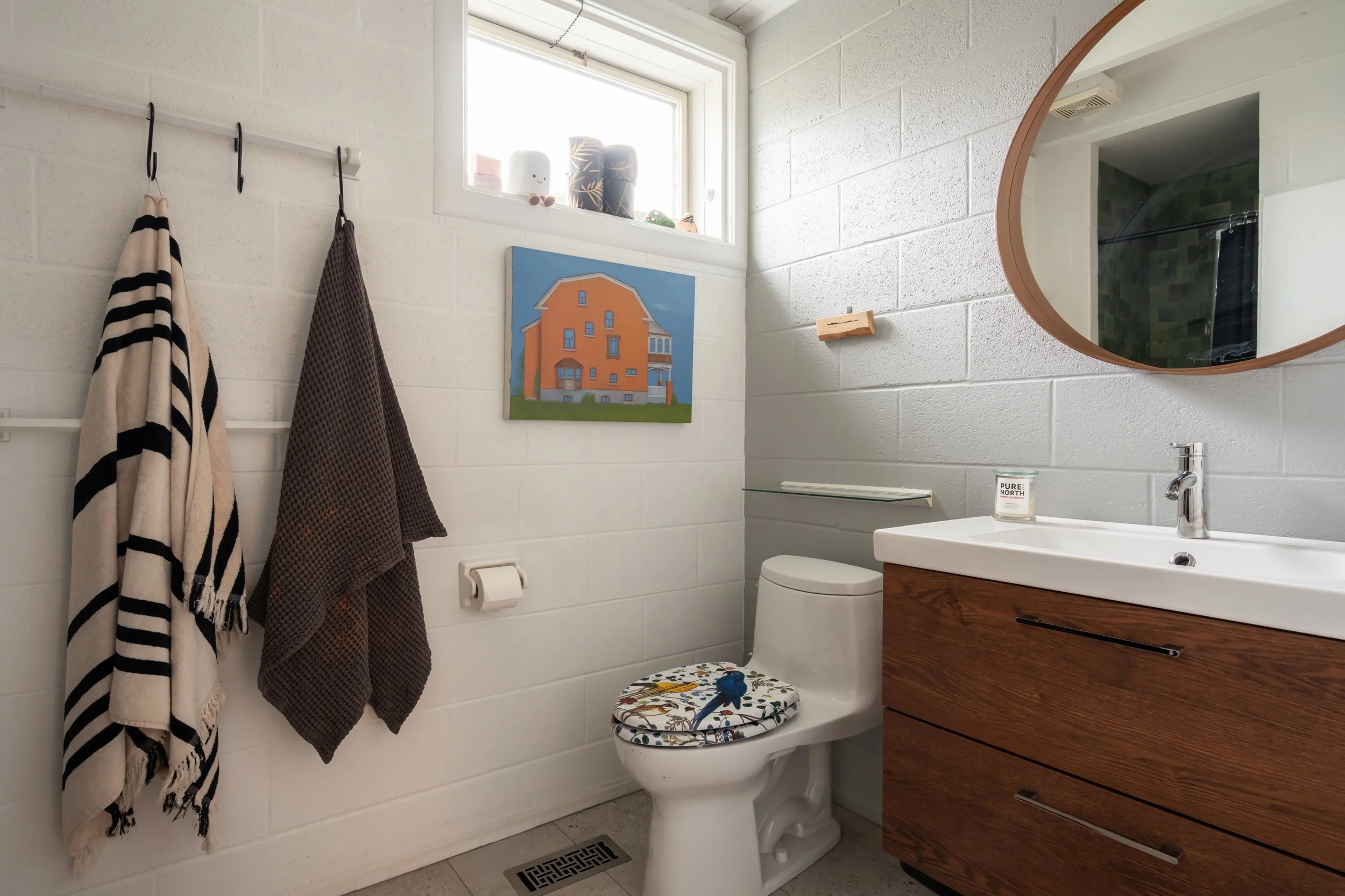 Bathroom with white brick walls, a wooden vanity with a white sink, a round mirror, a toilet with a decorative lid cover, a window, towels hanging on hooks, and artwork on the wall.