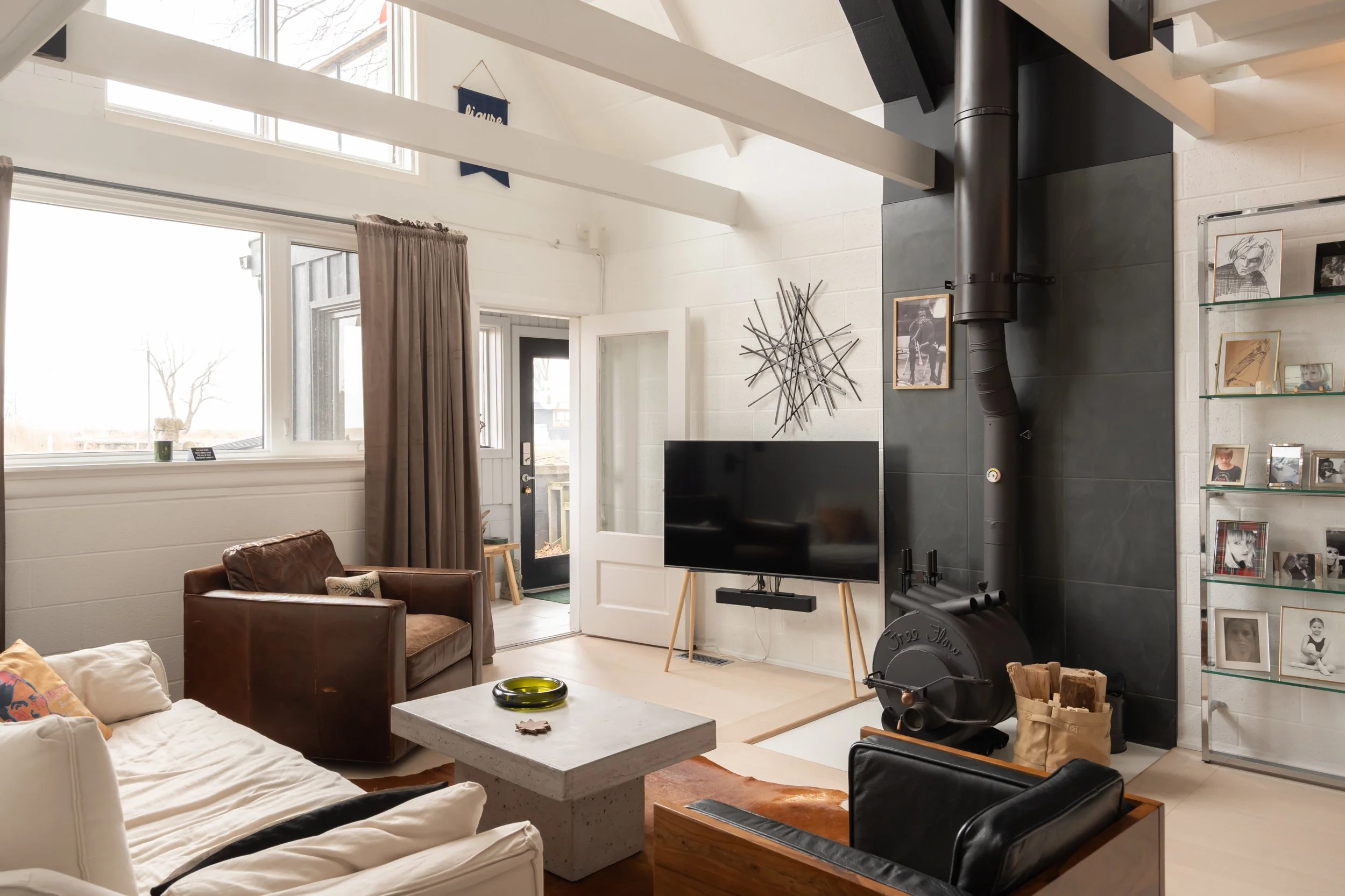 Living room with large windows, beige curtains, white walls, exposed ceiling beams, black tiled fireplace, black stove, mounted flat-screen TV, white coffee table, brown leather armchair, beige sofa with pillows, black armchair, glass display shelf w