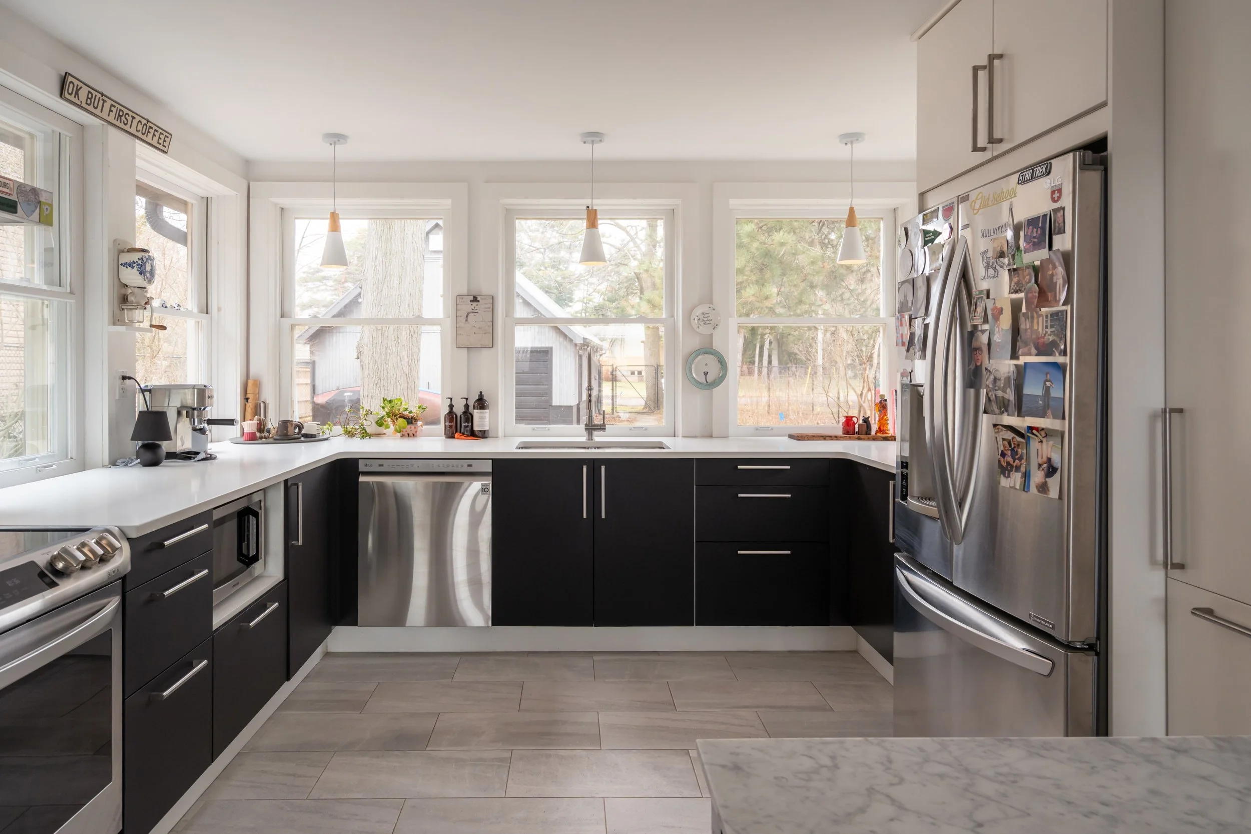 Modern kitchen with black and white cabinets, stainless steel appliances, large windows, pendant lights, and various photographs and magnets on the refrigerator.