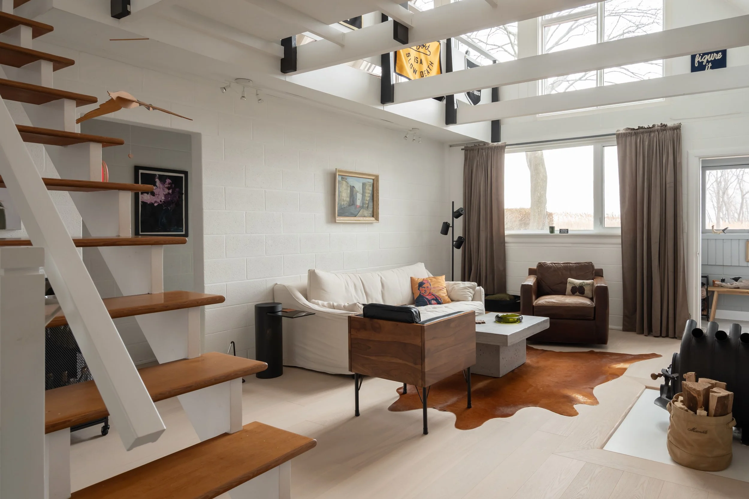 Living room with white brick wall, white sofa, brown leather chair, large windows with beige curtains, wooden staircase, cowhide rug, and chess set in front of fireplace.