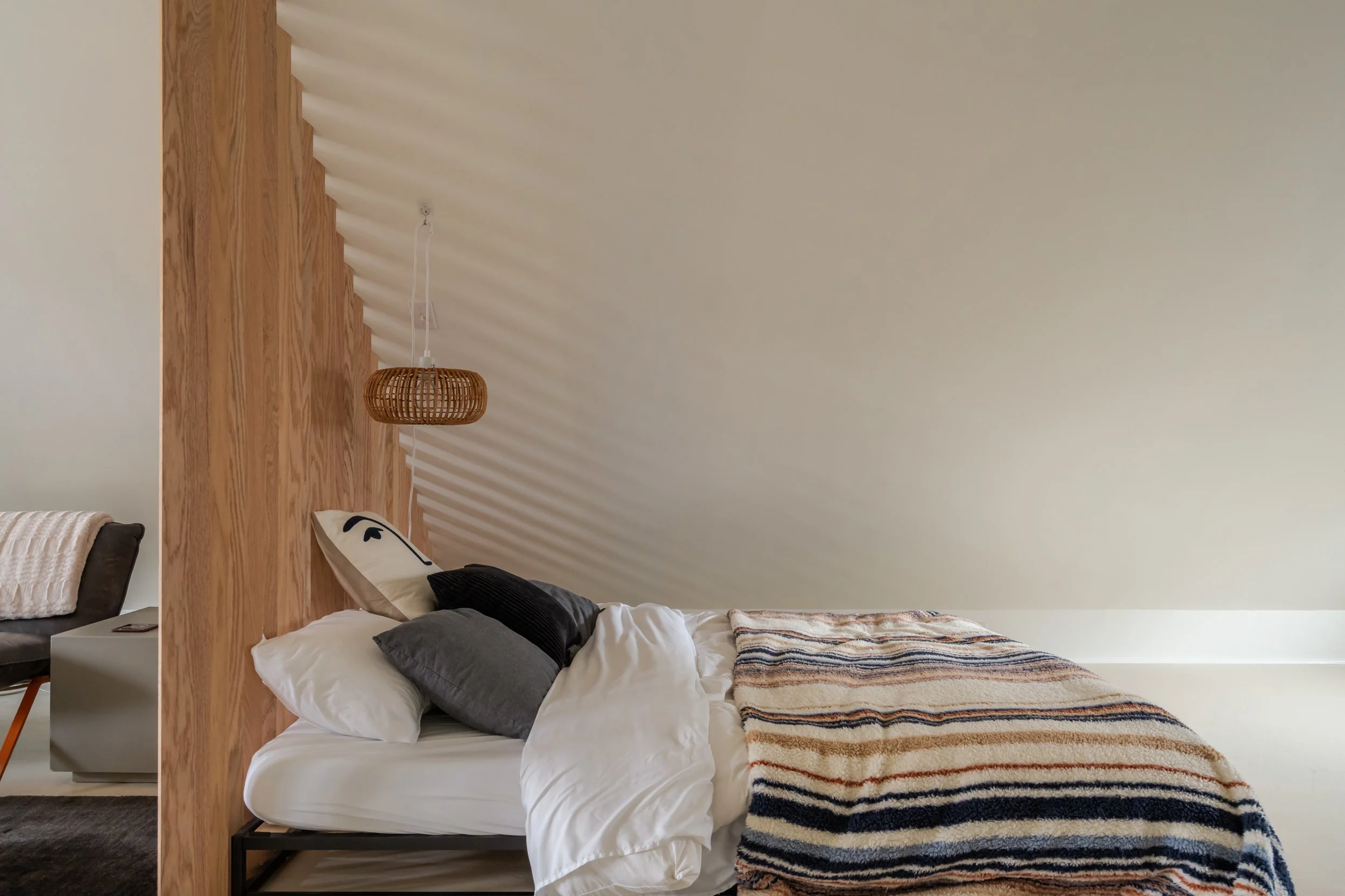 Bedroom with a sloped ceiling, a bed with white and gray bedding and pillows, and a striped blanket at the foot of the bed. A wooden headboard and a rattan pendant light are also visible.
