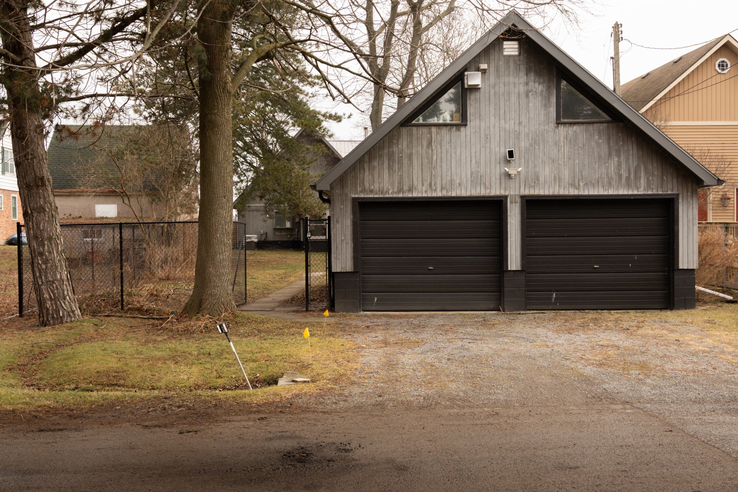A two-car garage with a wooden exterior painted grey and black garage doors, situated on a grassy lawn with trees and a neighborhood in the background.