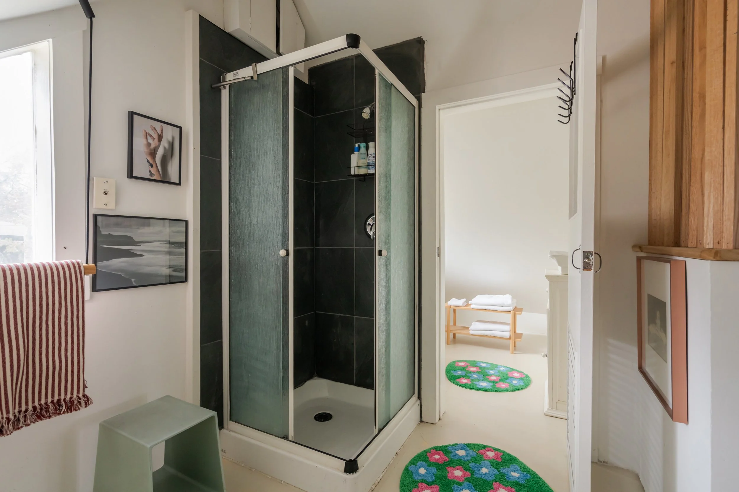 Bathroom with a walk-in shower with frosted glass doors, black tile walls, two colorful floral rugs on the floor, framed wall art, towel rack with red and white striped towel, and a small stool.
