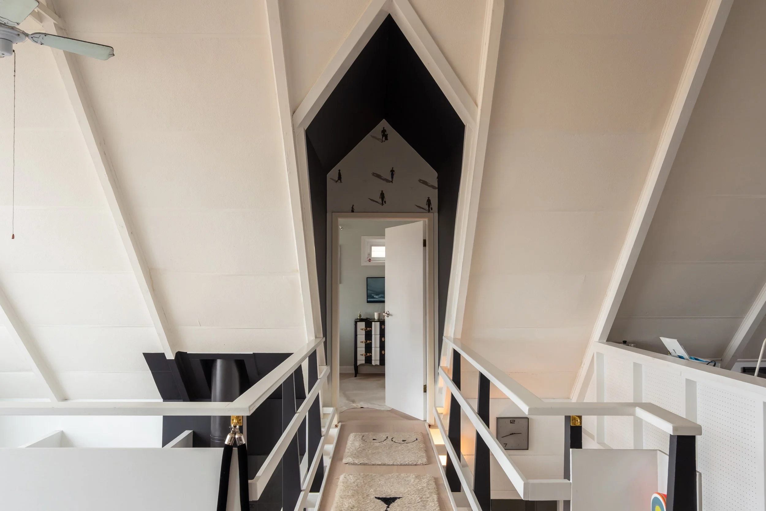 View of a hallway in an attic with white sloped ceilings, black and white railing, and a doorway leading to a room with a dresser, picture frame, and window.