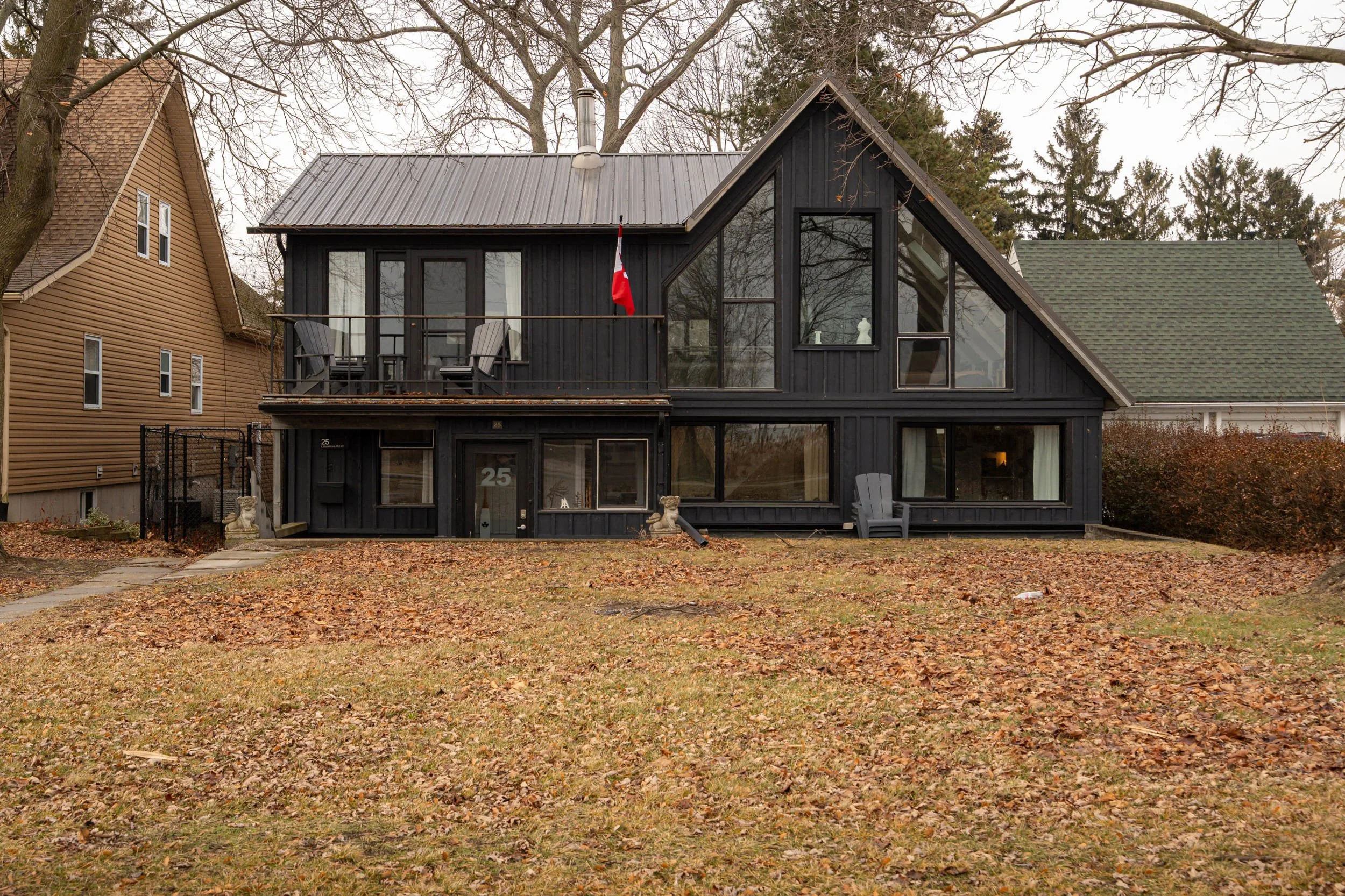 A two-story house painted black with large triangular windows on the upper floor. The house has a balcony with two chairs and a Canadian flag. The front yard has fallen leaves and some garden ornaments. Adjacent houses are visible on both sides, with